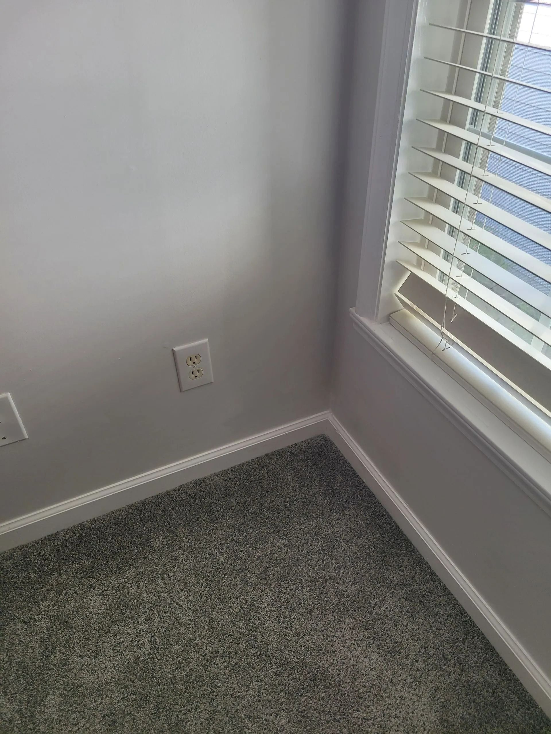 Empty carpeted corner with a window, white walls, baseboard, and electrical outlets.