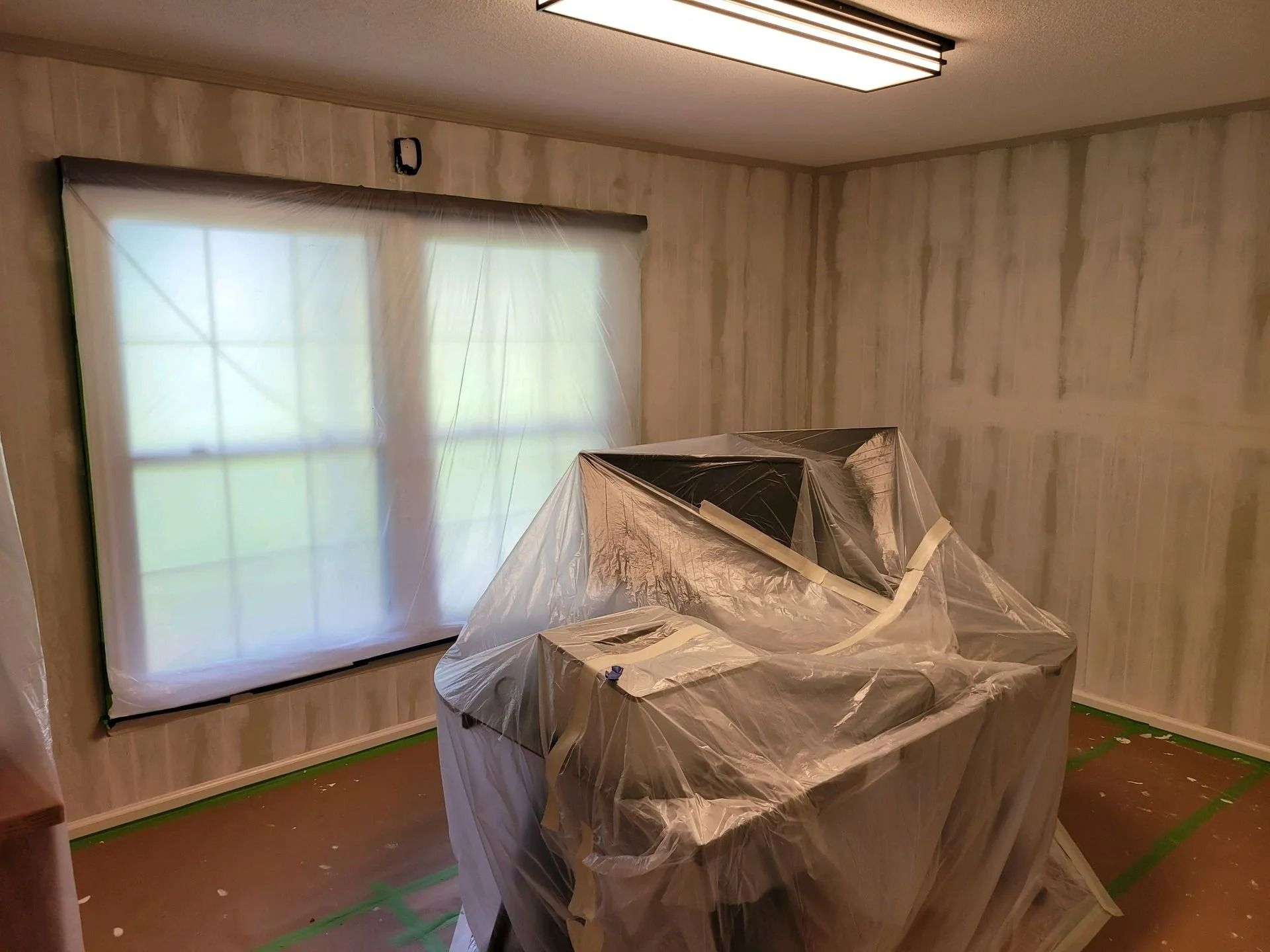 A room with a large window and a plastic-covered chair or machine on the floor, walls unfinished and taped off.