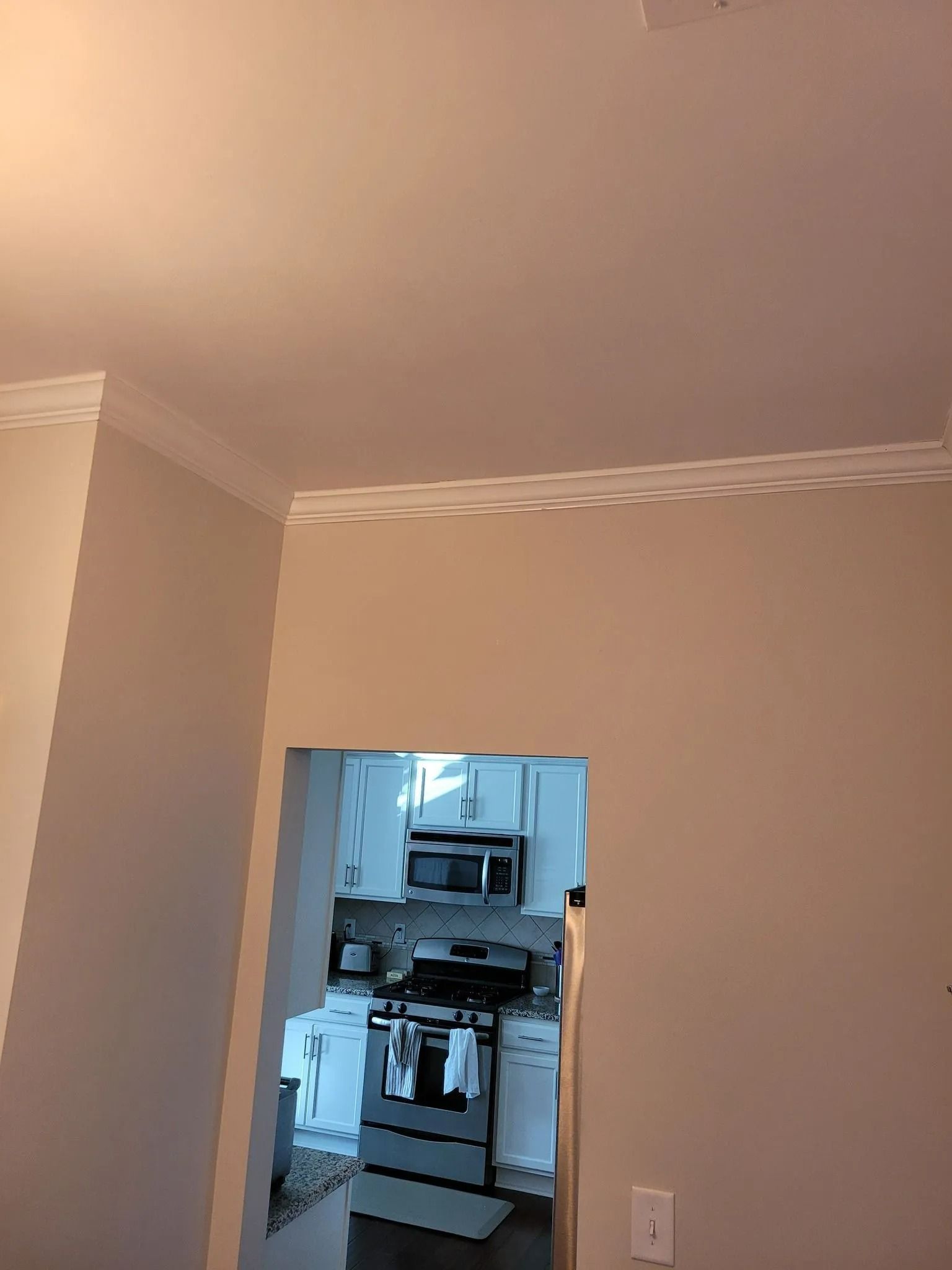 Empty beige room with crown molding, looking through a doorway into a small kitchen.