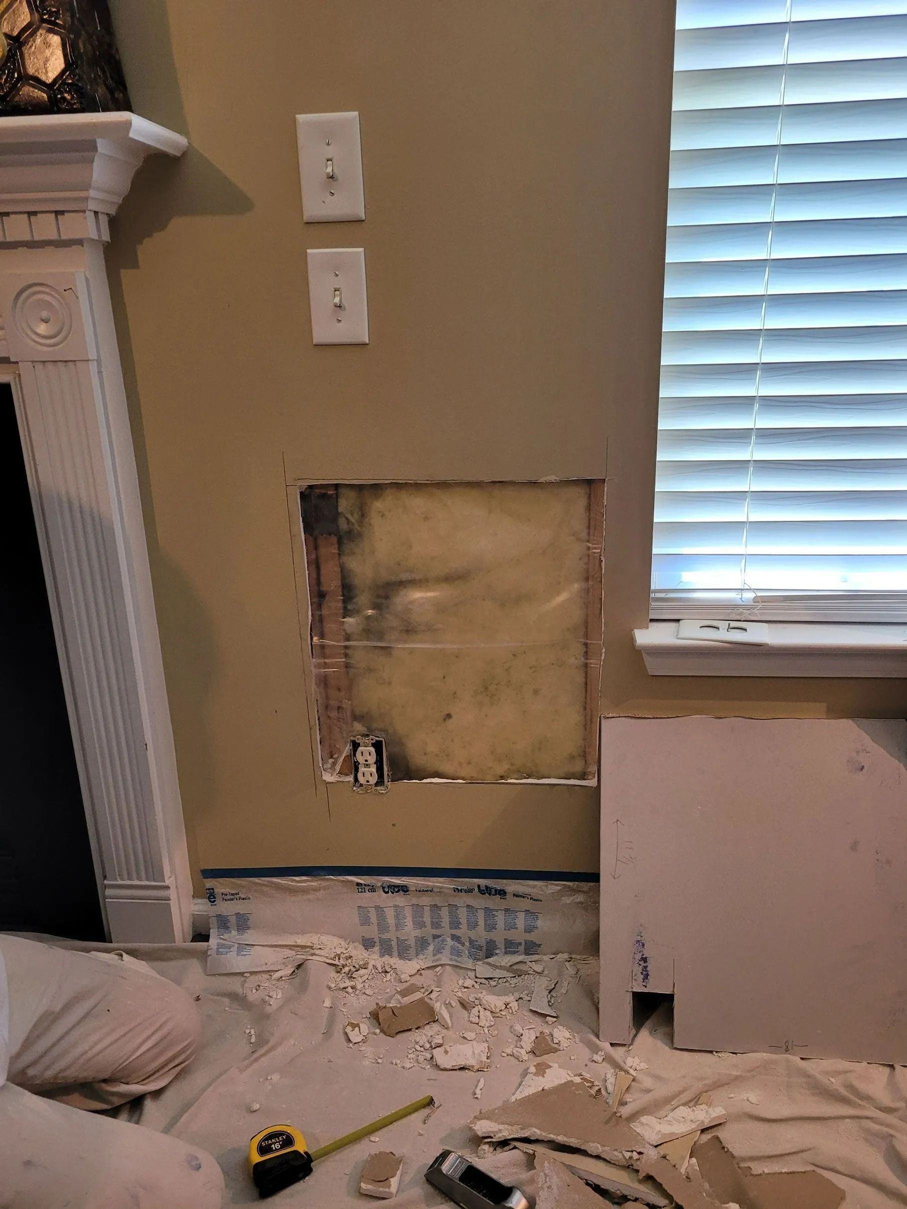 Damaged wall with exposed drywall, debris on floor, and a window with blinds in a room under repair