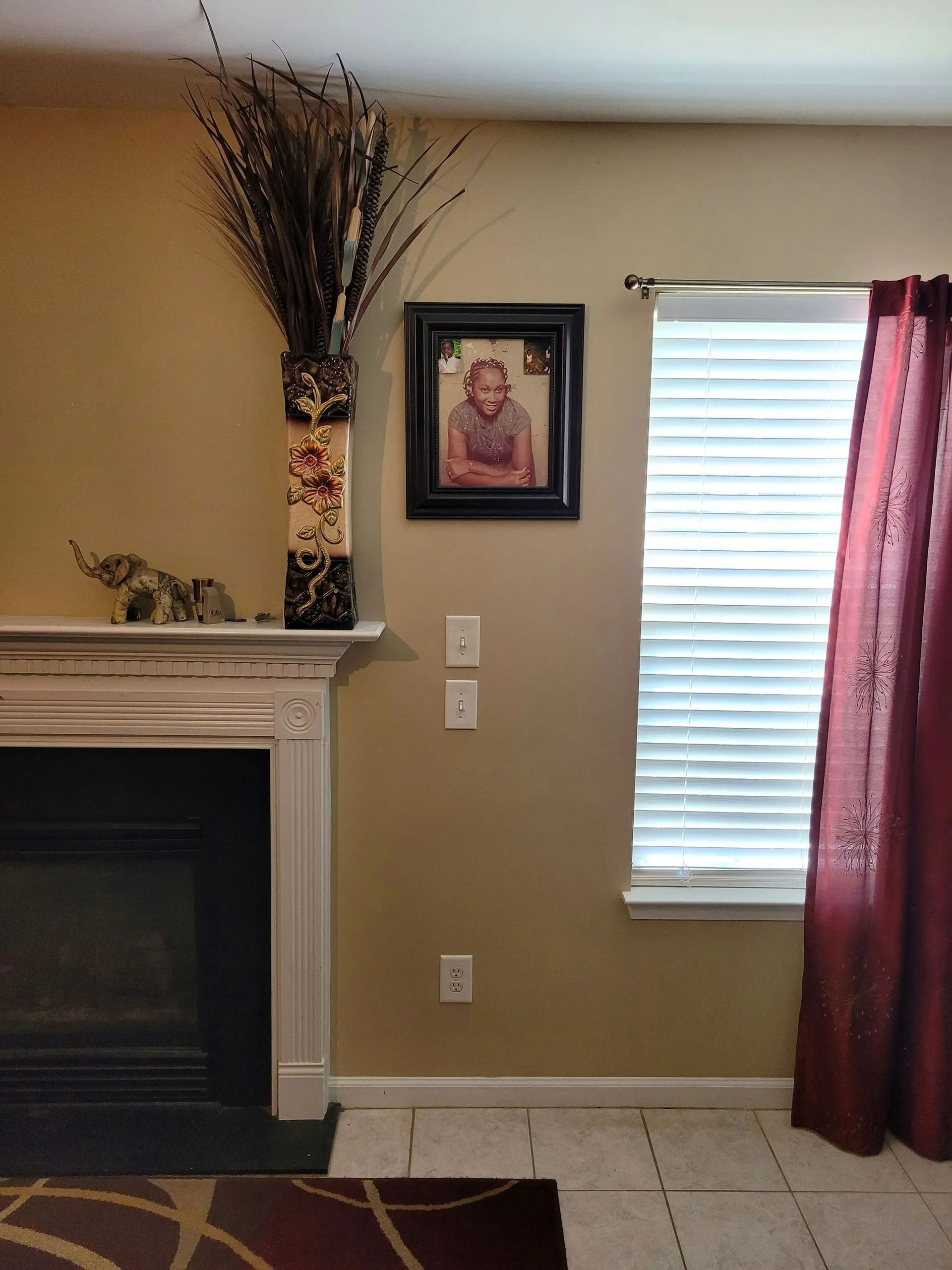 Living room with fireplace, framed portrait, tall dried floral arrangement, and window with burgundy curtains