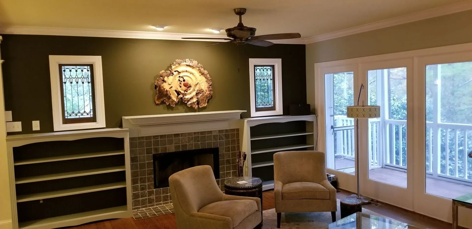 Living room with stone fireplace, built-in shelves, beige chairs, and large windows overlooking a deck