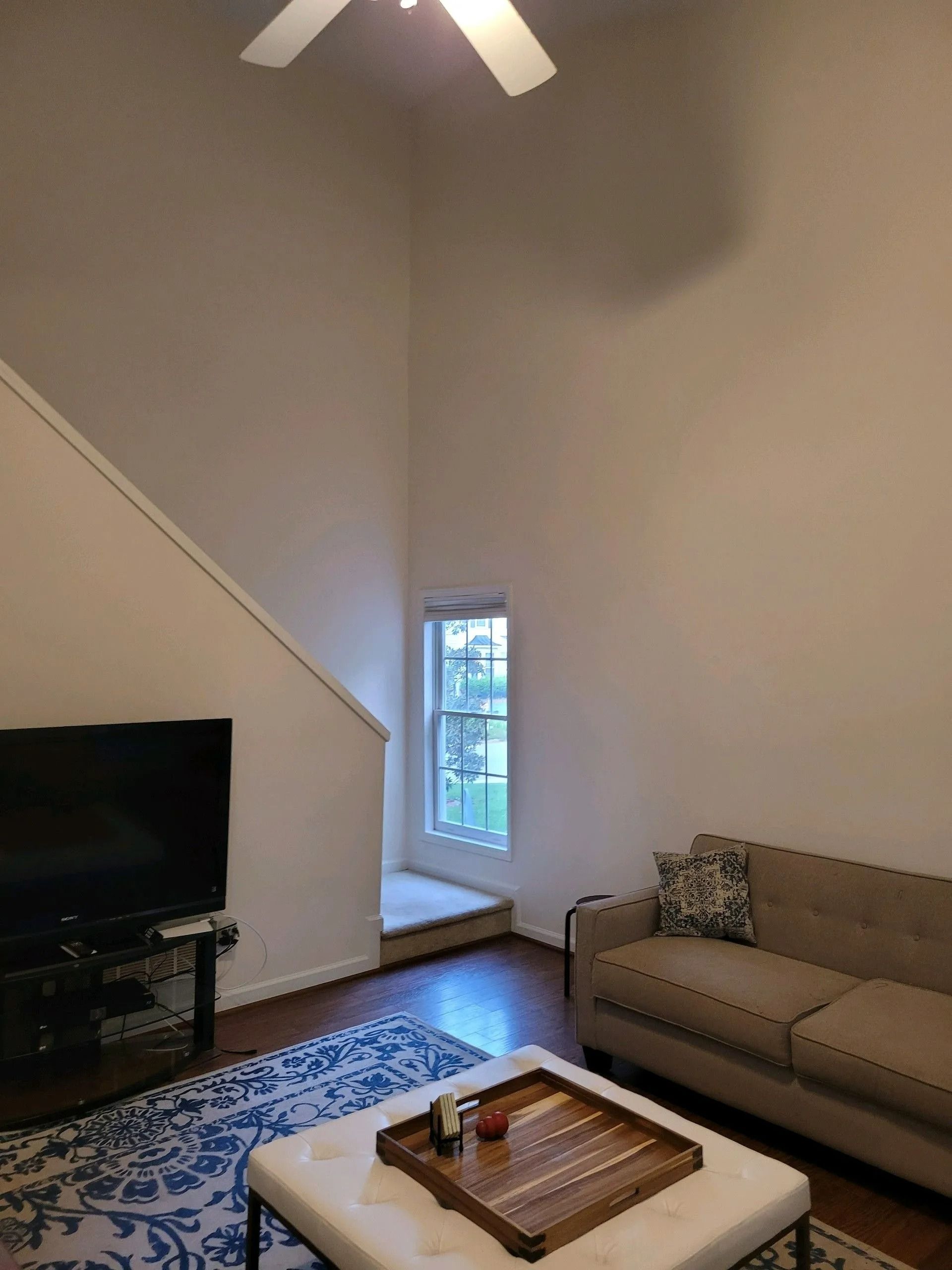 Living room with beige sofa, TV, patterned rug, coffee table, and high ceiling with ceiling fan.