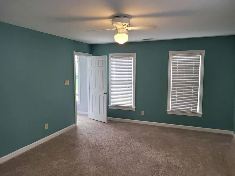 Empty teal bedroom with beige carpet, white door, two windows, and a ceiling fan-light.