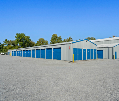 Storage units with blue doors, gray metal siding, and gravel parking lot under a blue sky.