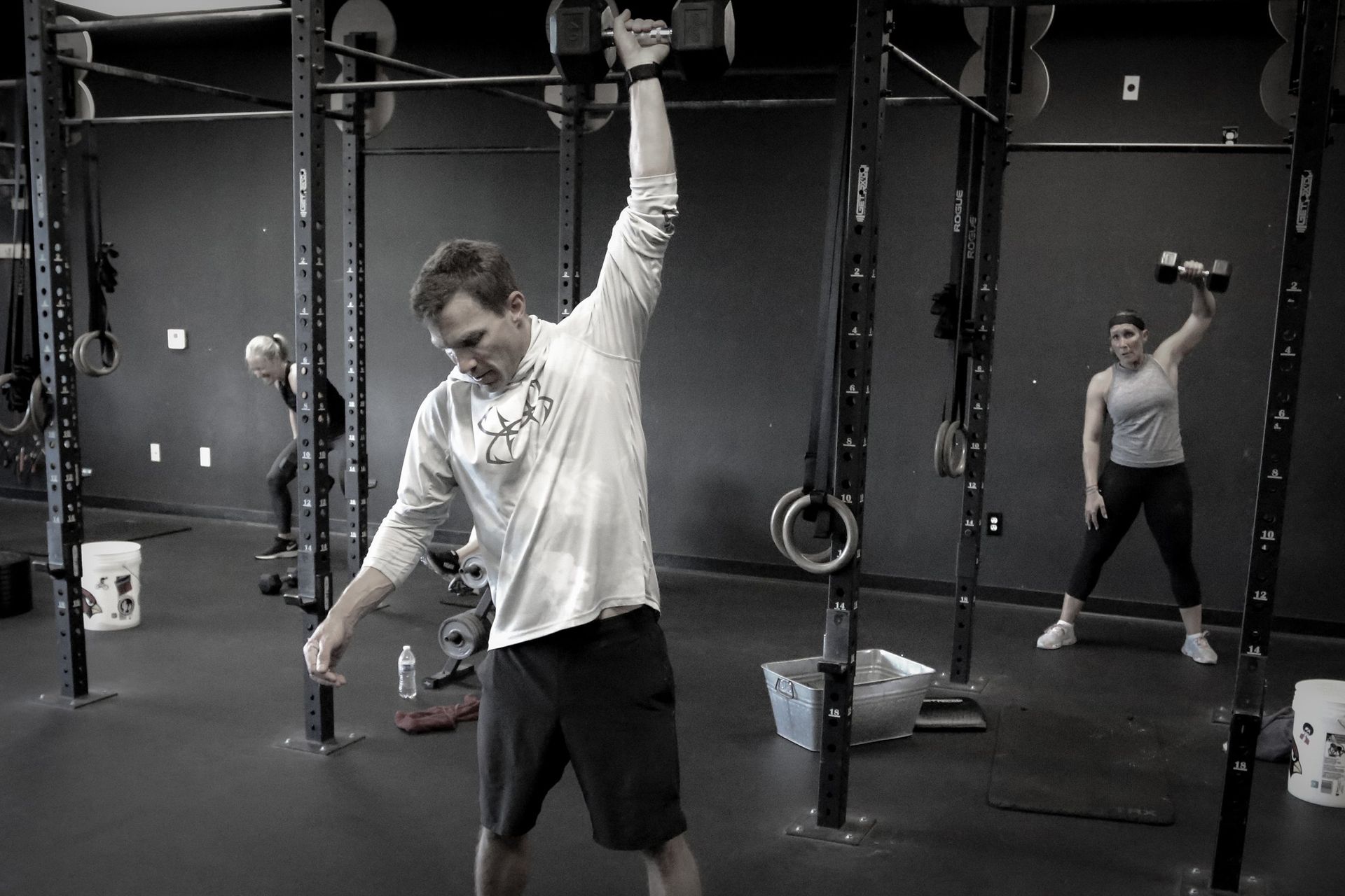 People exercising with kettlebells in a gym, one lifting overhead while another stands behind.