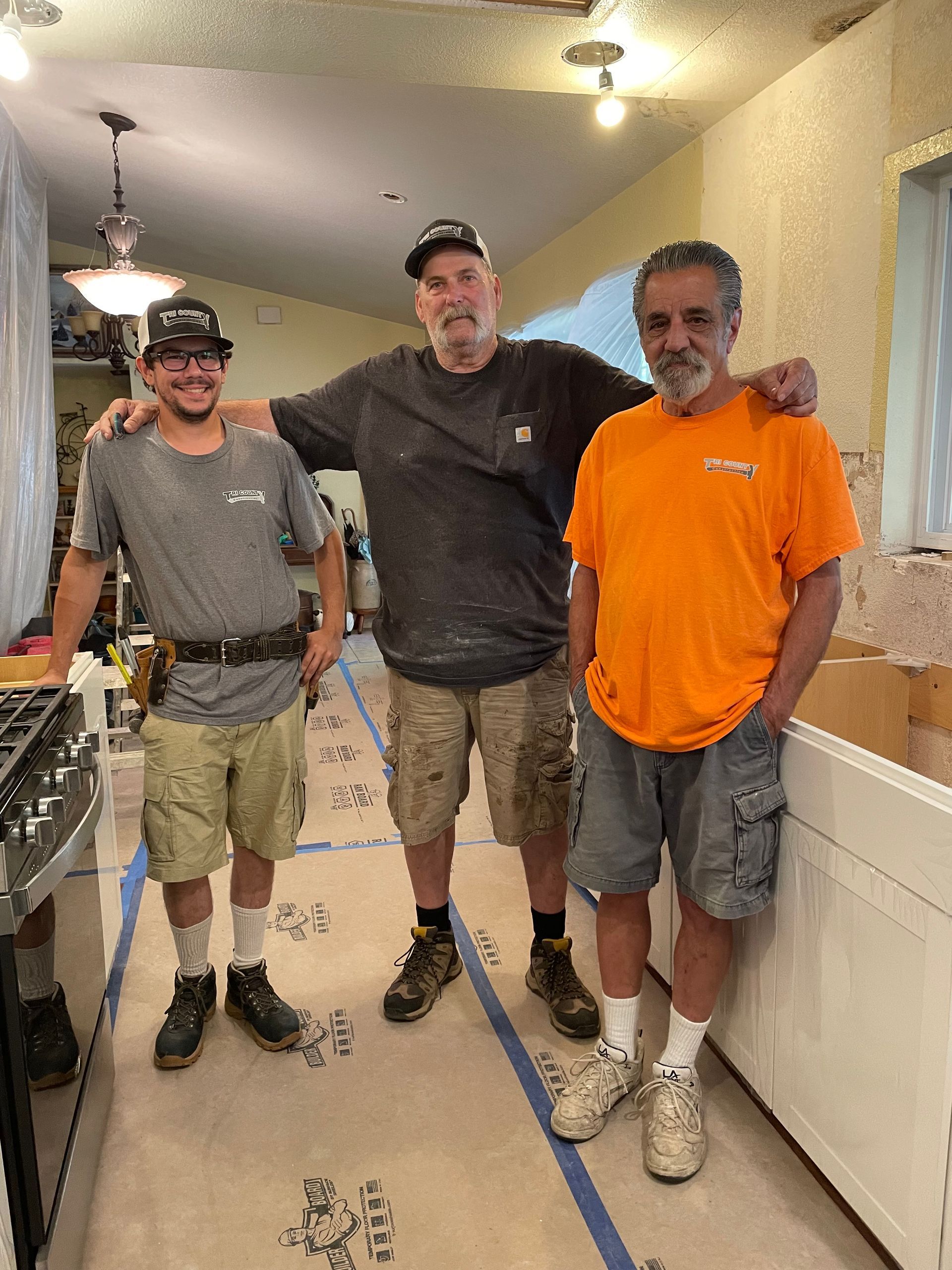Three men standing together in a narrow kitchen hallway, smiling, with one in an orange shirt and another on a counter.