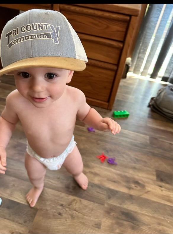 Toddler standing indoors on a wood floor, wearing a gray cap and diaper, with toys scattered nearby.