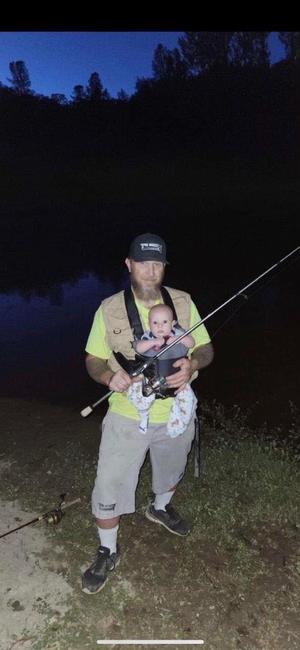 Person standing outdoors at dusk, holding a fishing rod and gear on a grassy path.