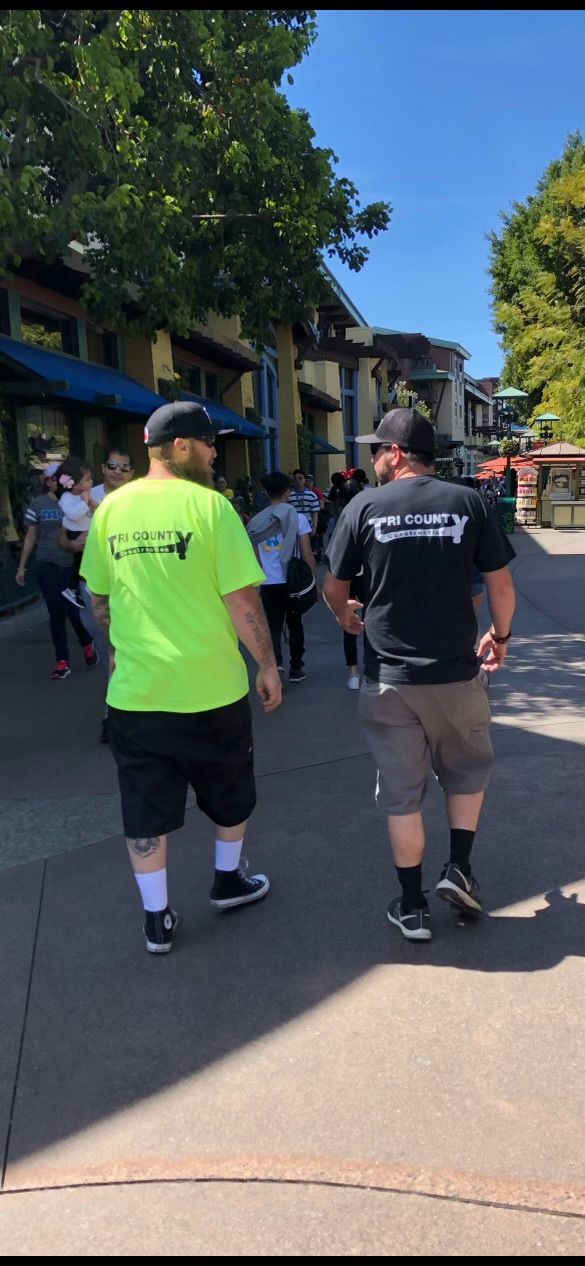 Two people walking on a sunny sidewalk near shops and trees, one in a neon green shirt.