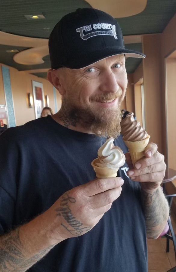 Man smiling and holding two soft-serve ice cream cones indoors, wearing a black cap and T-shirt