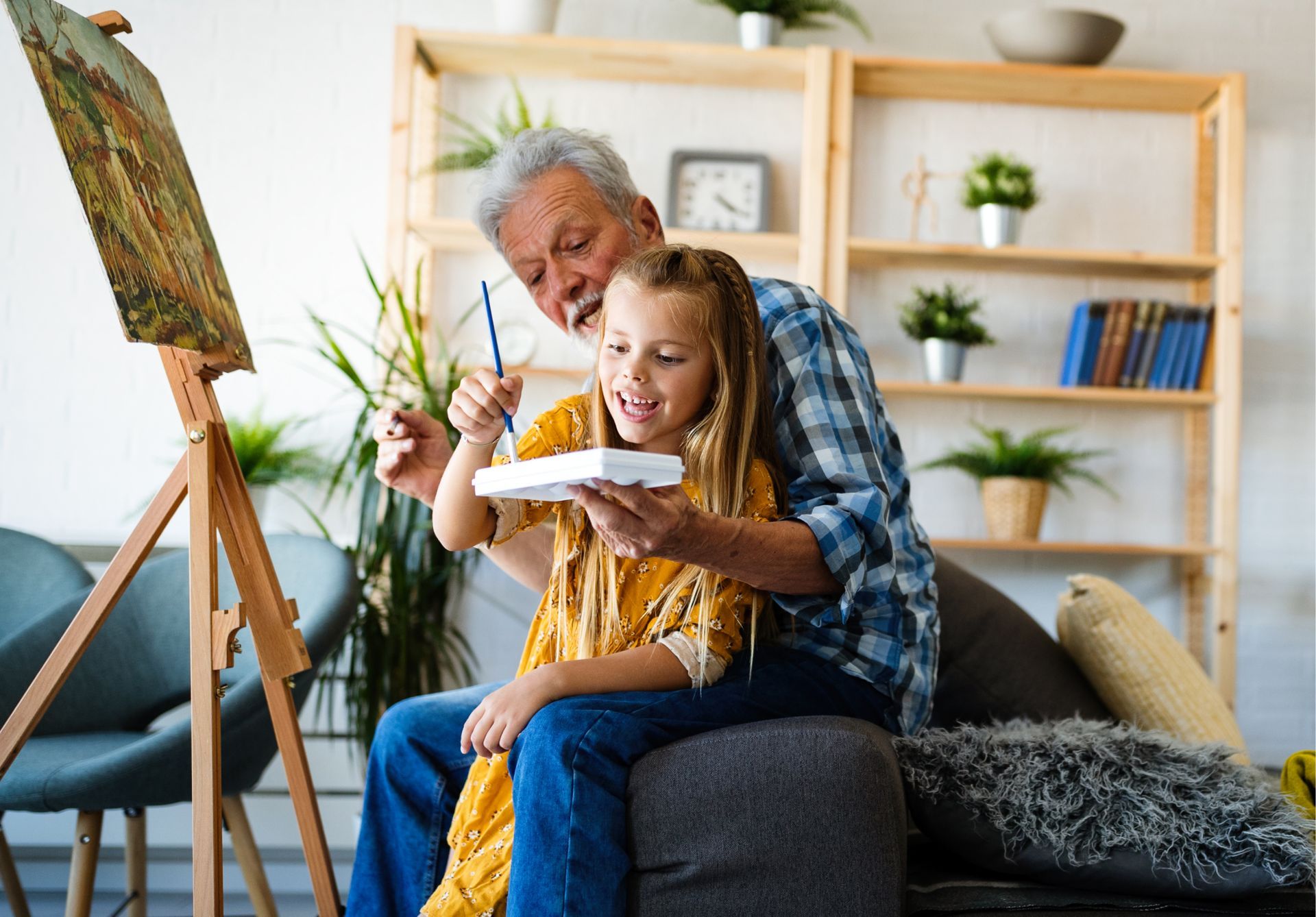 An elderly man is teaching a little girl how to paint on an easel.