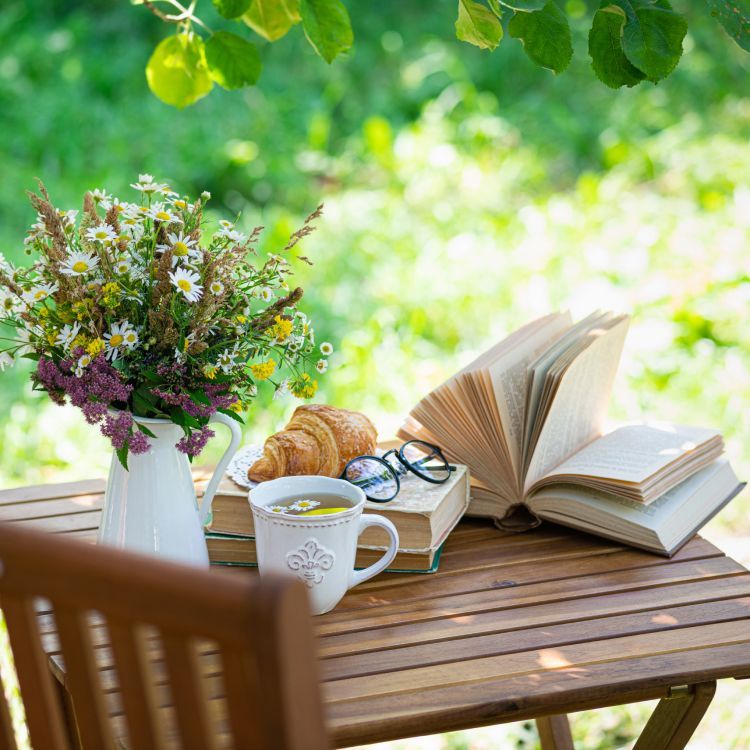 A wooden table with books , a cup of tea , a croissant and a vase of flowers.