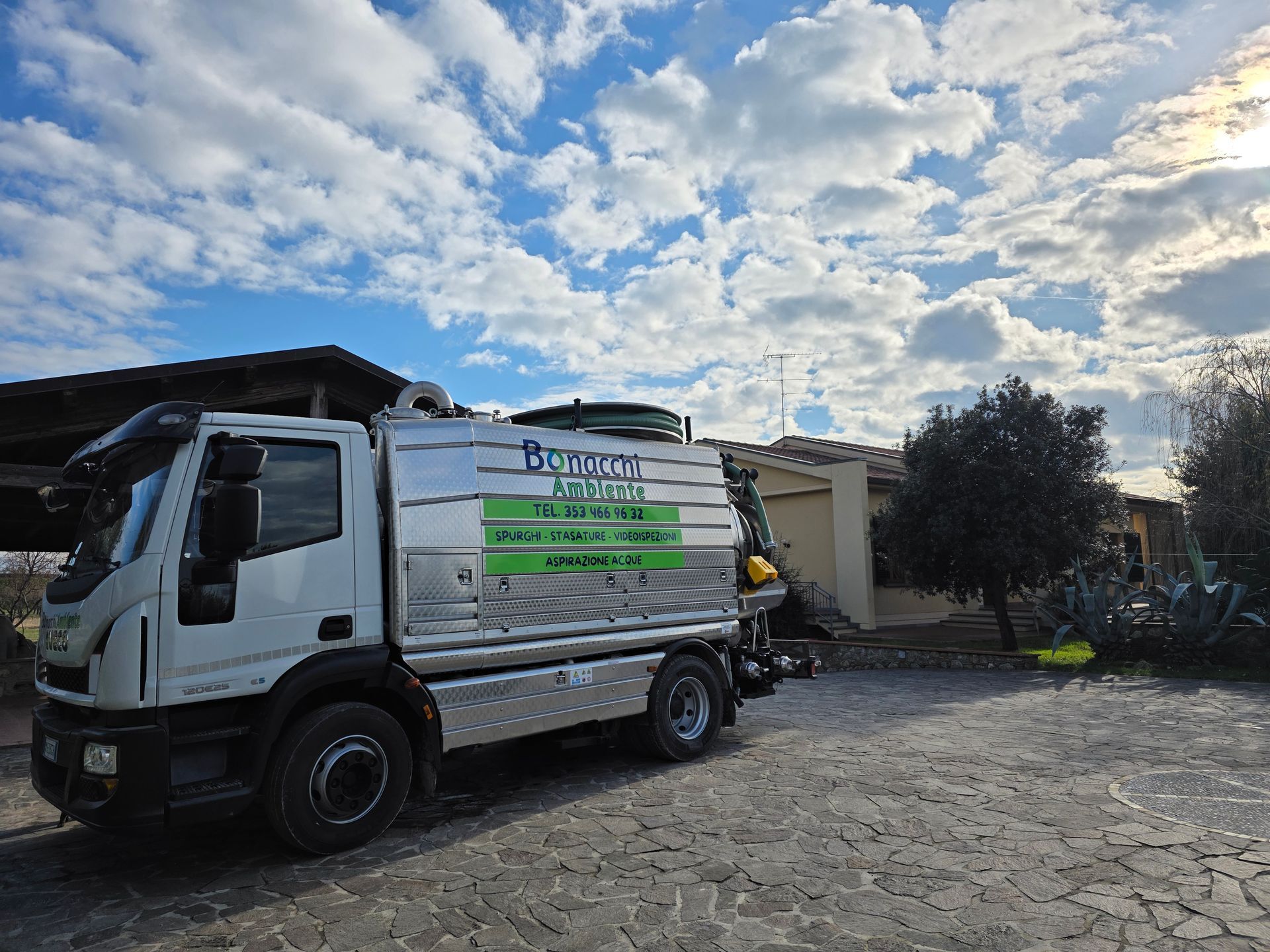 Un furgone bianco per le consegne con un cartello laterale verde è parcheggiato su un vialetto lastricato vicino a un edificio, sotto un cielo nuvoloso.