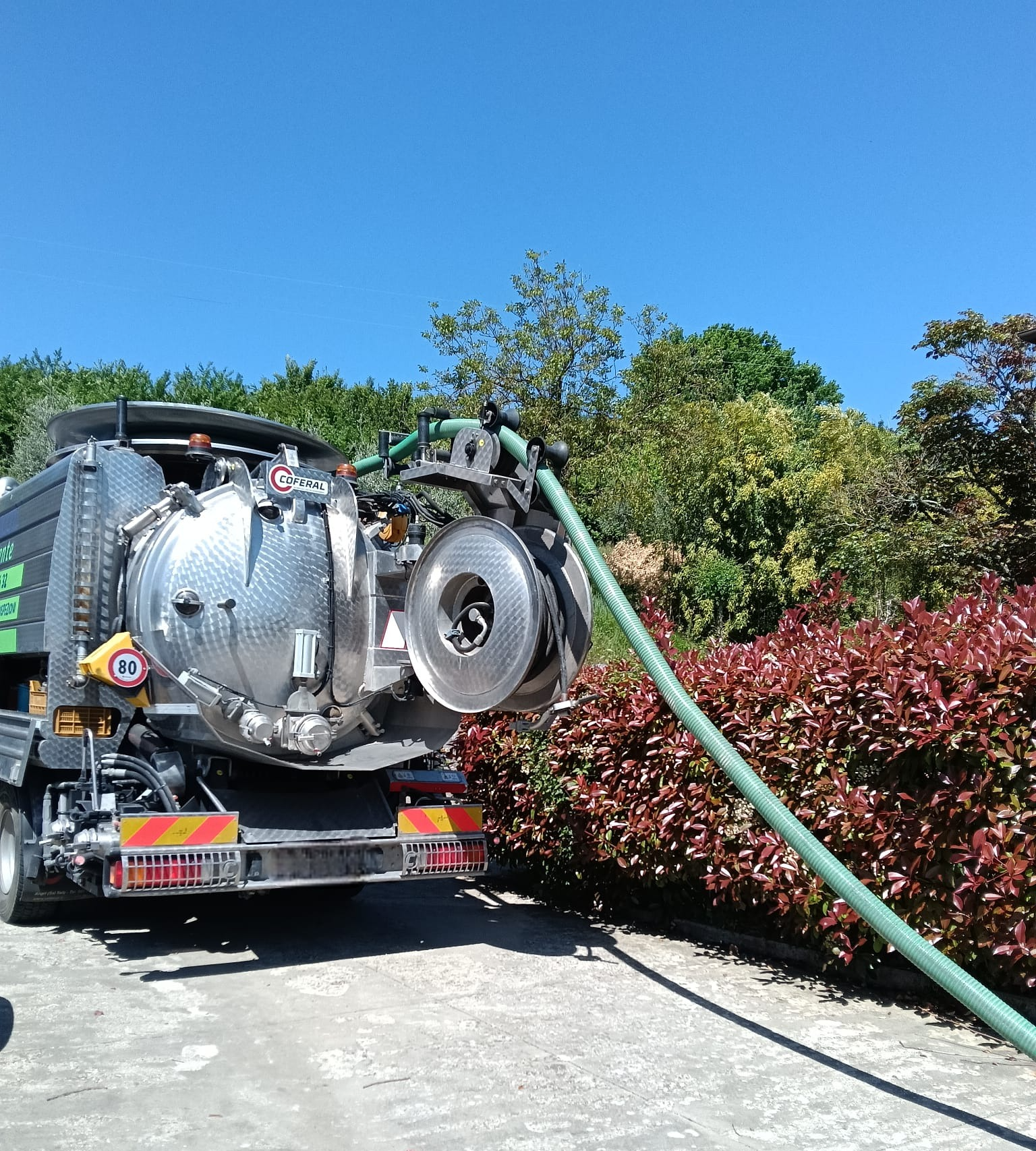 Camion aspiratore che pompa acqua da un tubo in un serbatoio in un vialetto soleggiato accanto a una siepe.
