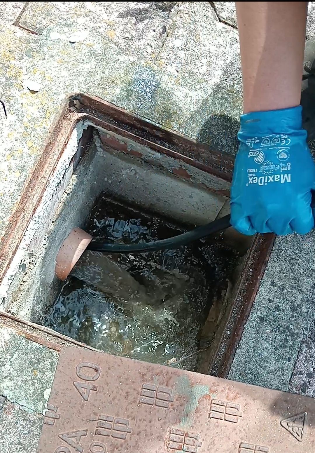 Mano guantata di blu che utilizza un cavo in un tombino aperto pieno d'acqua e detriti.