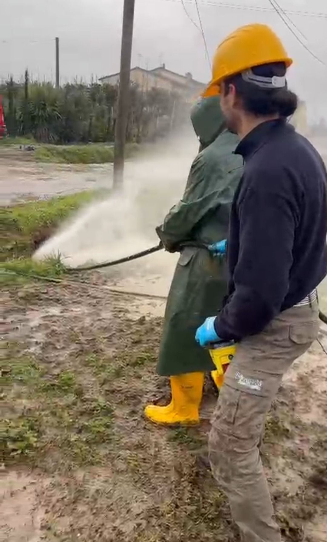 Due operai con indumenti antipioggia e stivali gialli usano un tubo per spruzzare acqua all'aperto.