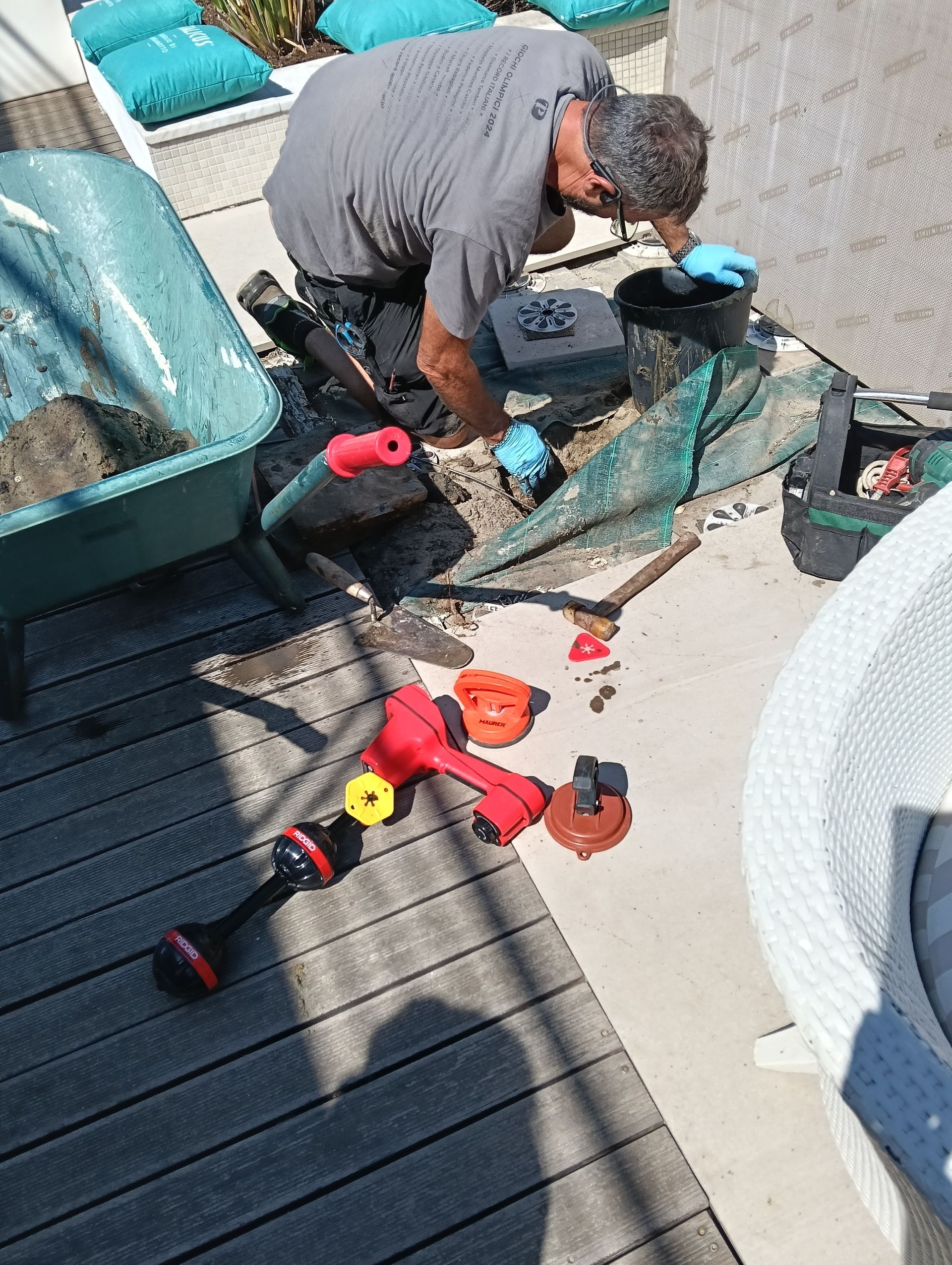 Una persona ripara una sedia da giardino a bordo piscina, con attrezzi e pezzi di ricambio sparsi sul terrazzo.