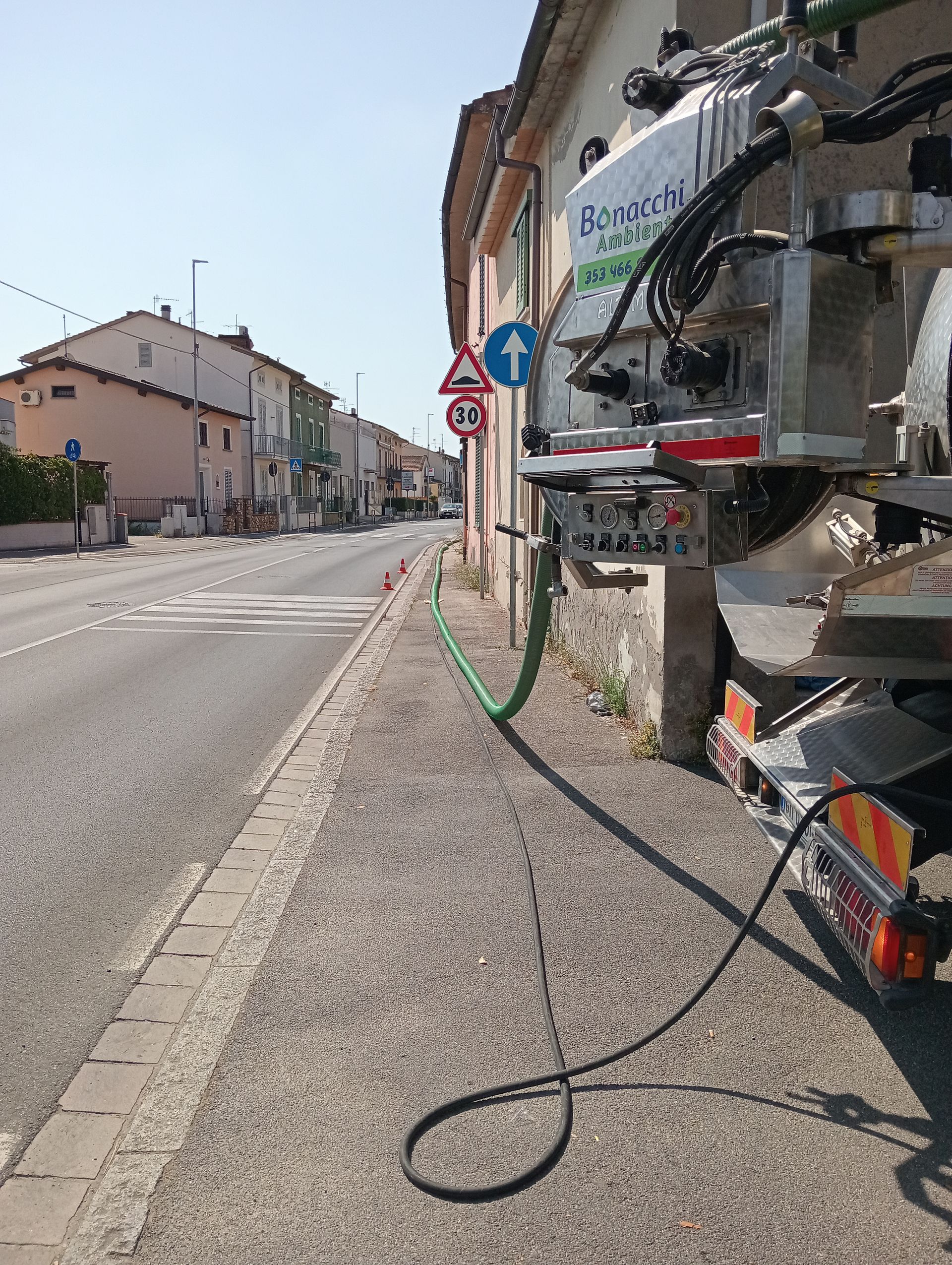 Veduta stradale con un grande veicolo da lavoro sulla destra e un tubo flessibile sul marciapiede in una tranquilla via di paese.
