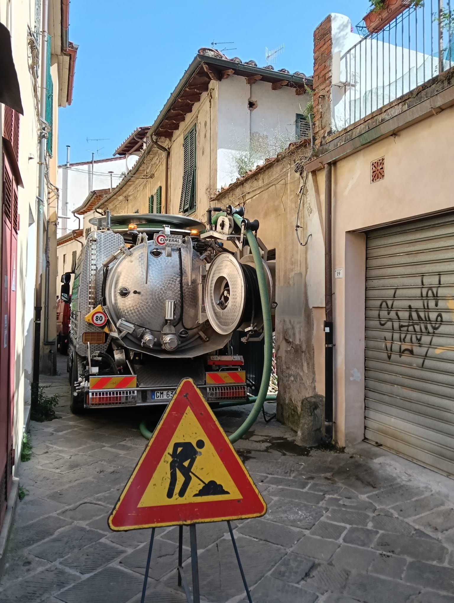 Un camion per la pulizia delle fognature in una strada stretta con un cartello di lavori stradali davanti.