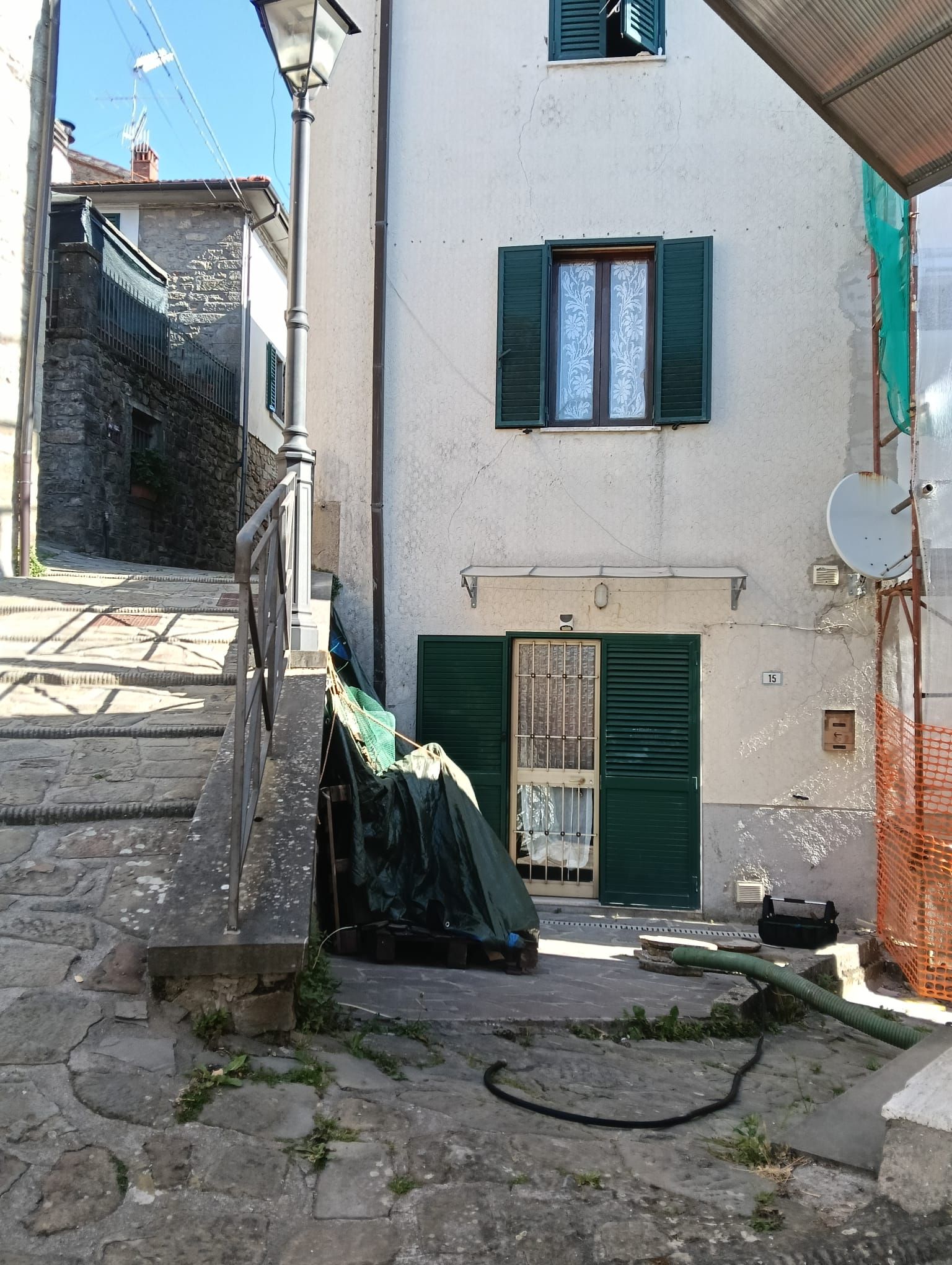 Stretto vicolo in pietra accanto a un edificio bianco con persiane verdi e una porta verde più bassa, in piena luce del sole.