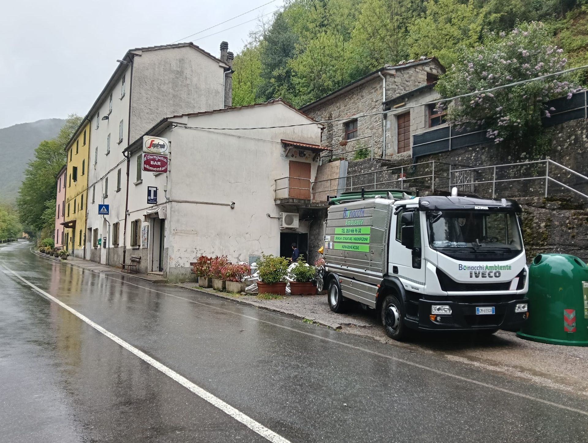 Via di paese sotto la pioggia, con un camioncino bianco di servizio accanto a edifici in pietra e vasi di fiori.