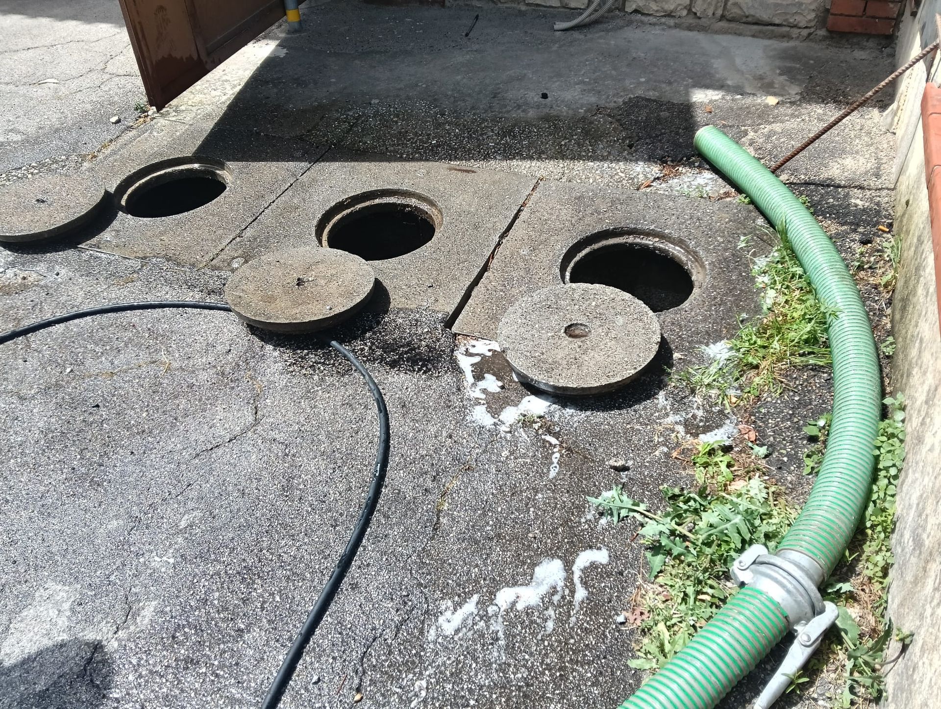 Due tombini aperti su una pavimentazione irregolare, accanto a un tubo verde e un cavo nero vicino a un muro di mattoni.
