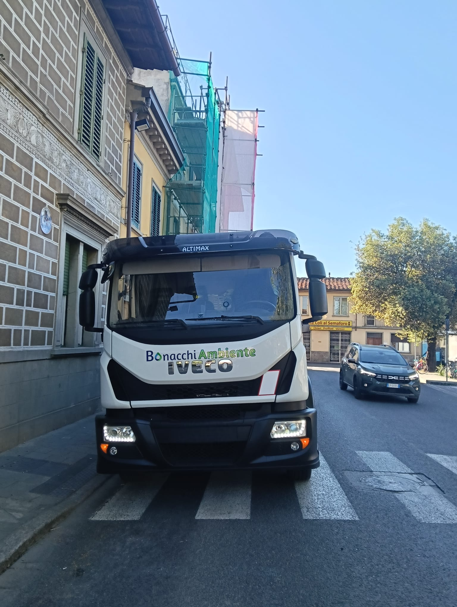 Un camioncino bianco per le consegne è parcheggiato in una strada stretta accanto a un edificio in pietra e a un'impalcatura, con delle auto sullo sfondo.
