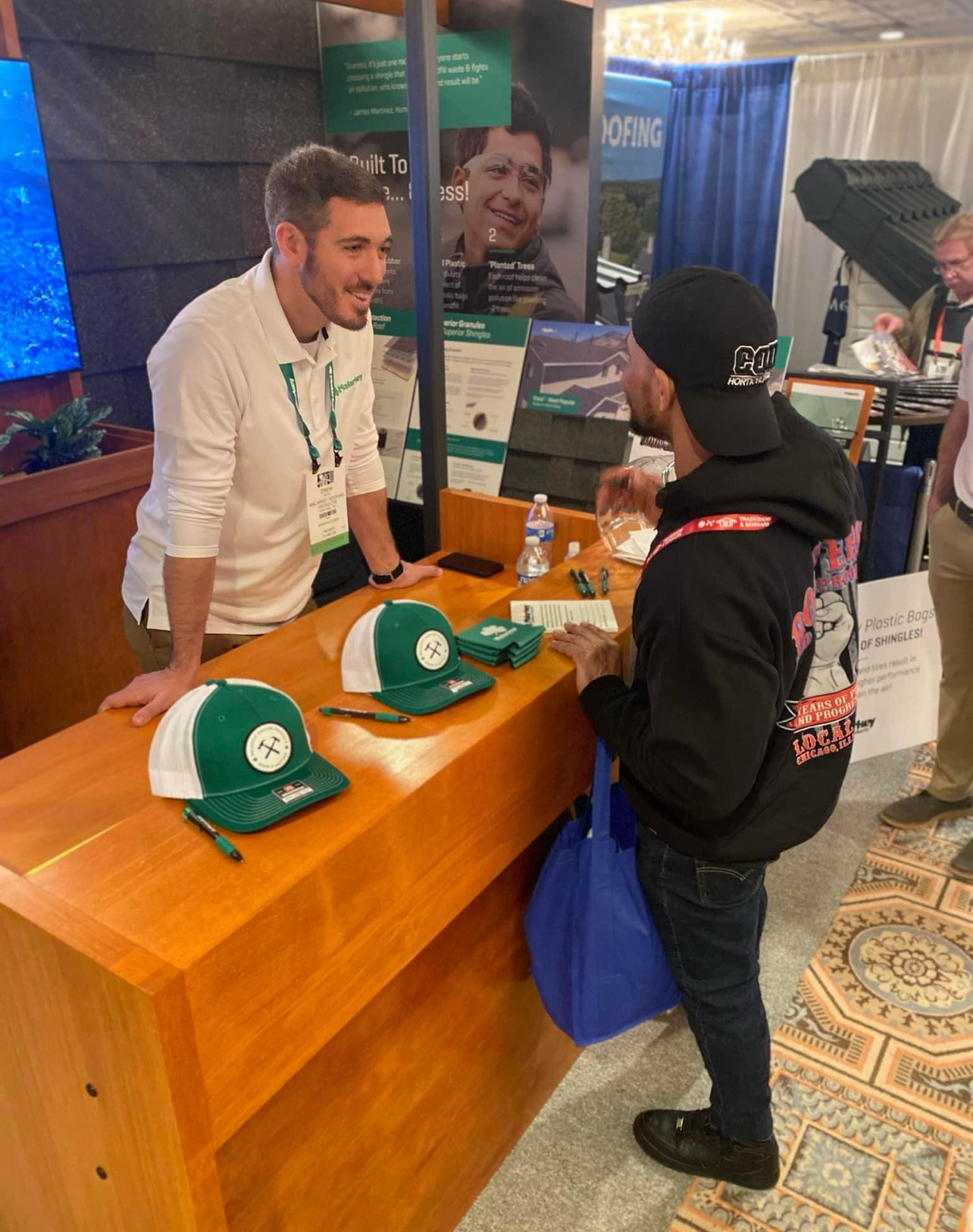 A man is talking to another man at a table with hats on it