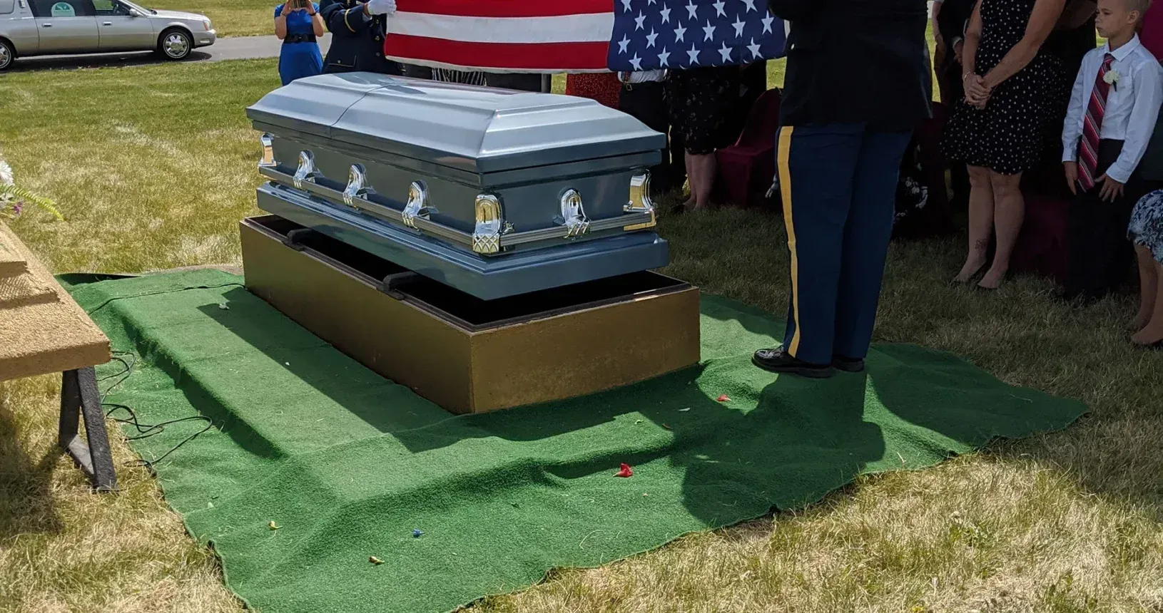 Small silver casket on a stand at an outdoor memorial, with mourners standing nearby and a U.S. flag draped behind.