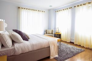 A bedroom with a bed featuring purple and white pillows, a shaggy rug, and windows with sheer curtains.