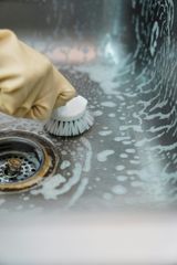 A gloved hand uses a circular white scrub brush to clean a stainless steel kitchen sink covered in soap suds.