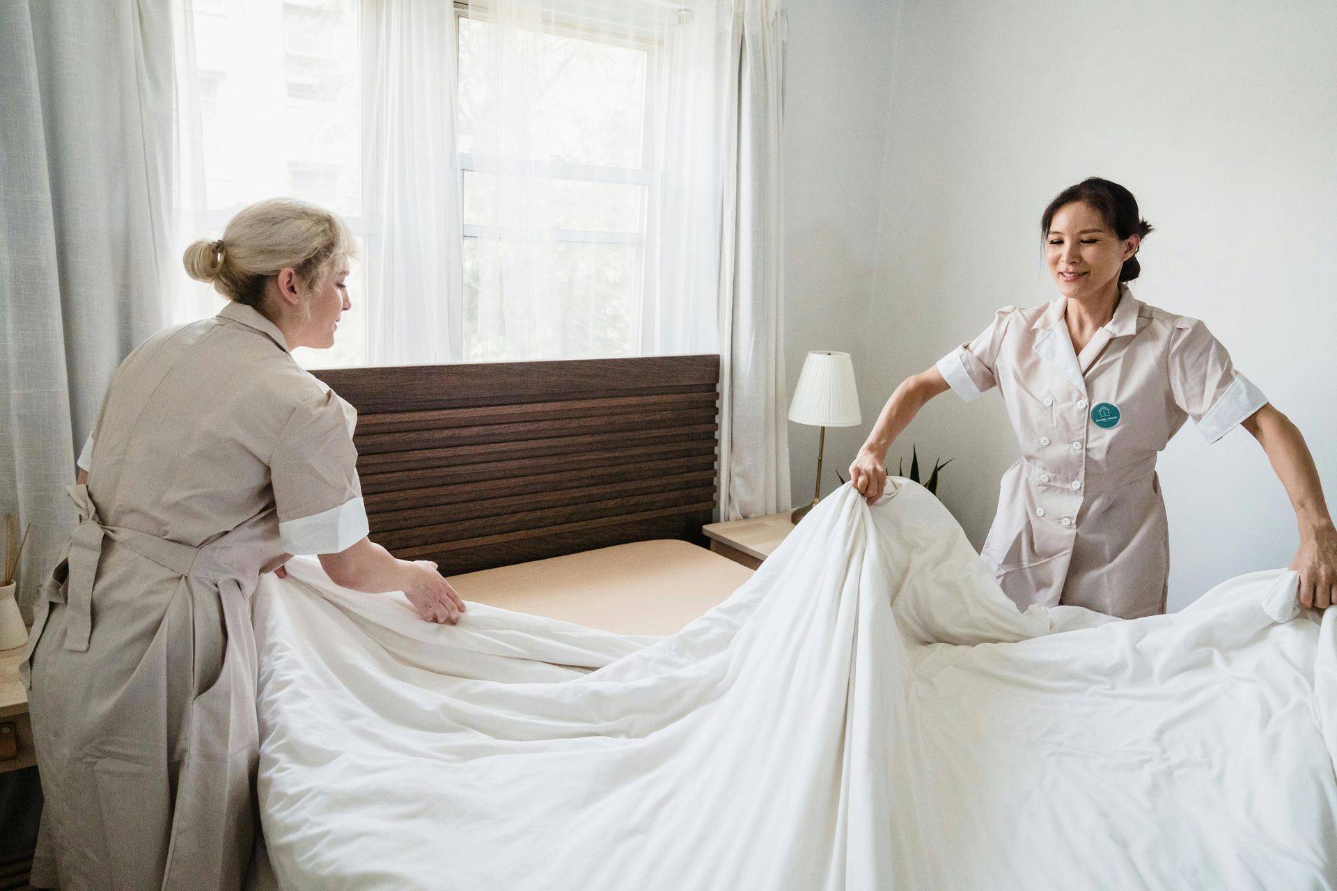 Two hotel housekeepers in uniform work together to spread a fresh white sheet over a made-up bed in a bright room.