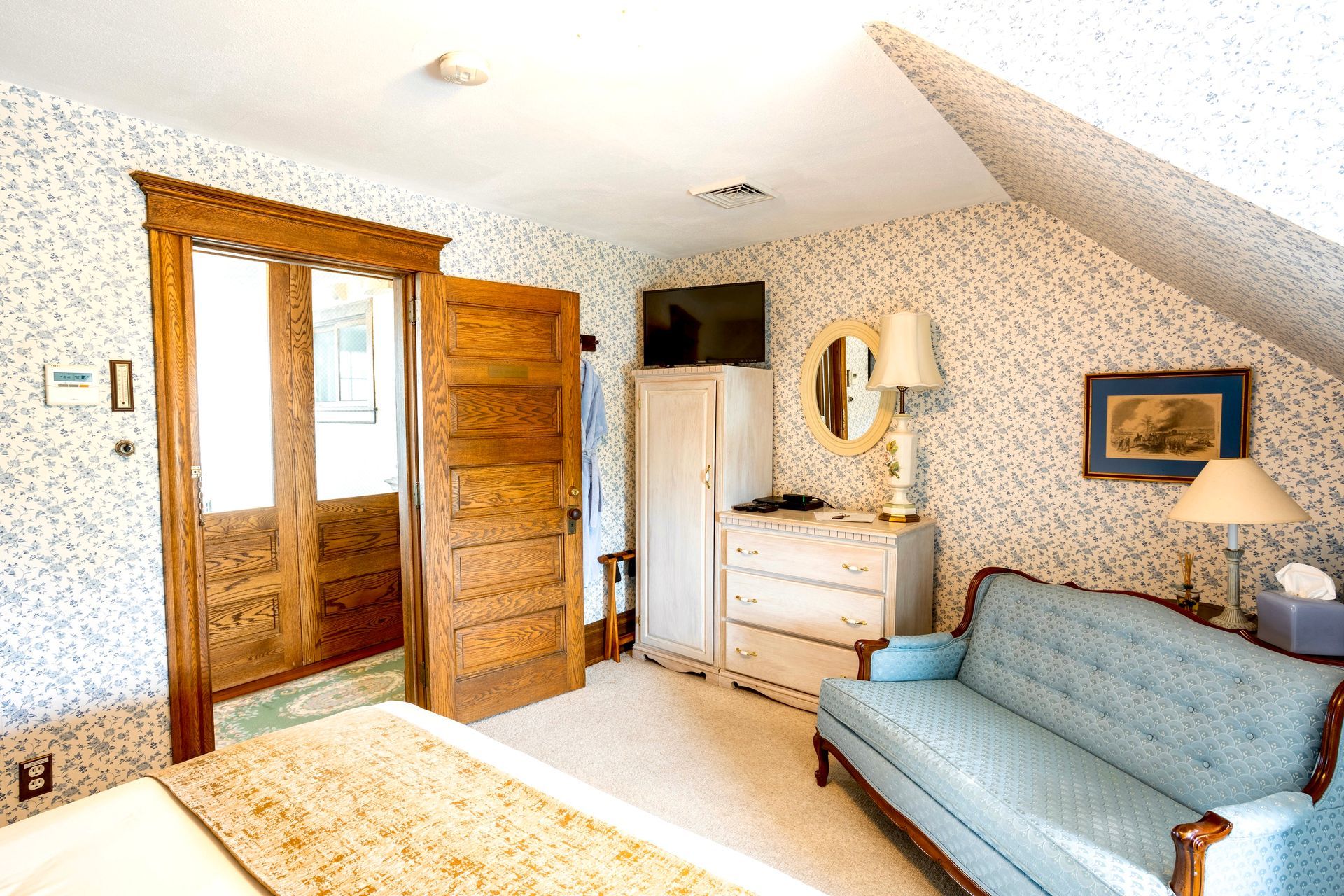 A cozy bedroom with floral wallpaper, wood paneling, a blue sofa, white dresser, and a table lamp under a sloped ceiling.