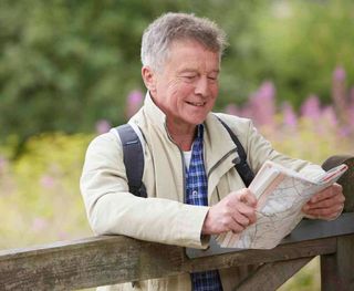 A man is leaning on a wooden fence reading a map.