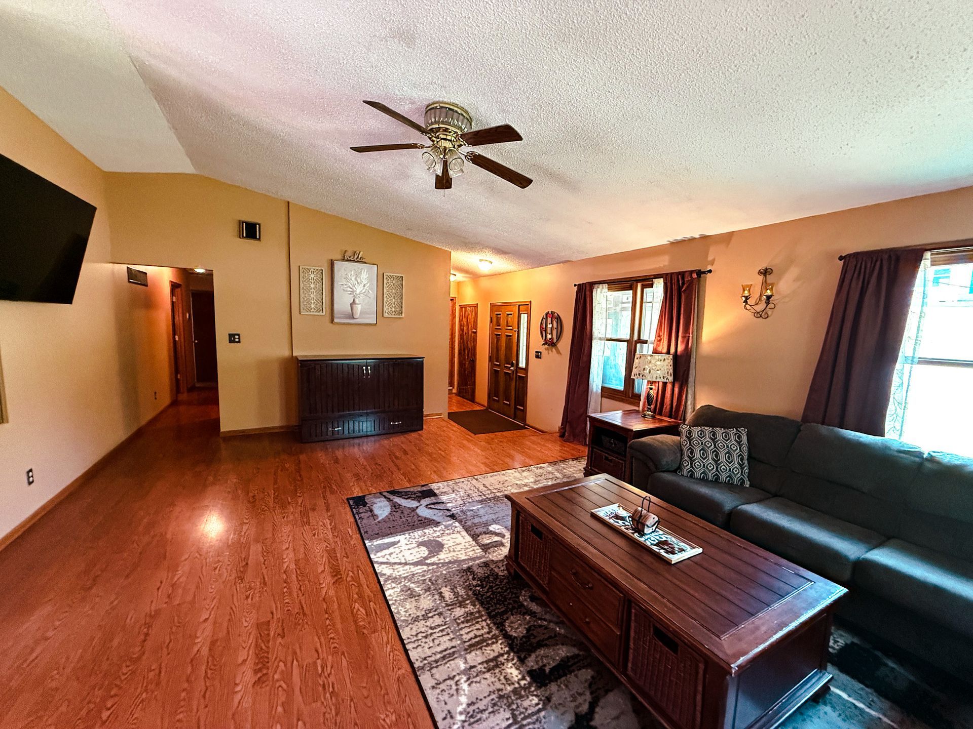 Living room with hardwood floors, dark sofa, coffee table, ceiling fan, and large windows with curtains