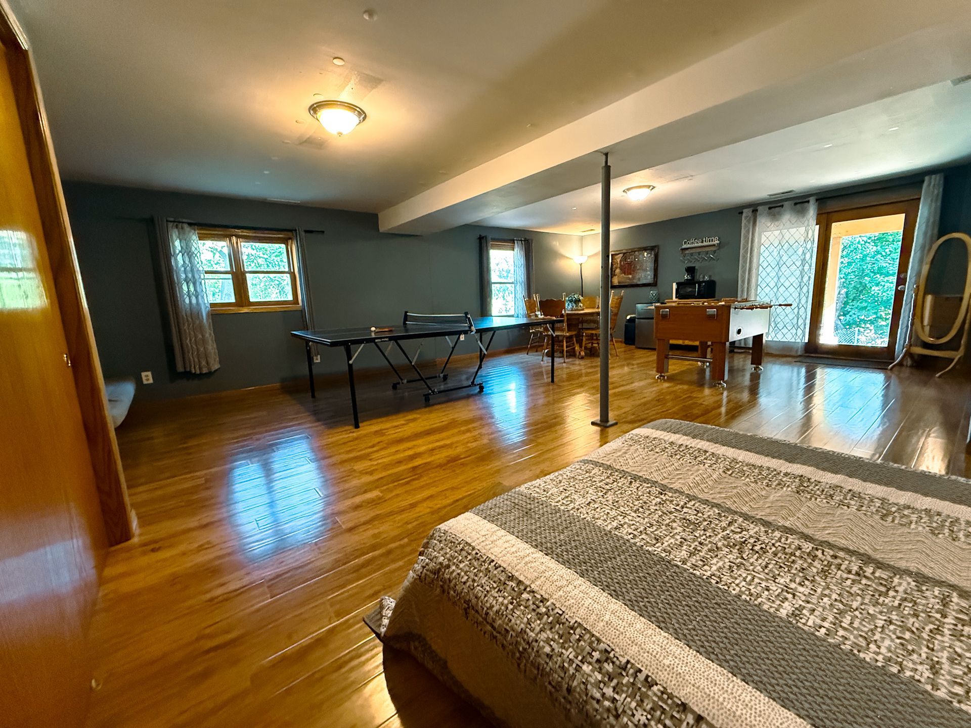 Spacious room with wood floors, bed in foreground, ping-pong table, desk, and large windows.