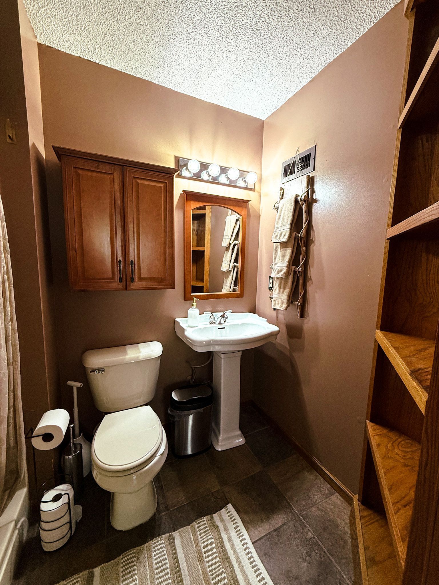 Compact tan bathroom with toilet, pedestal sink, wood cabinet, towel rack, and shelving.