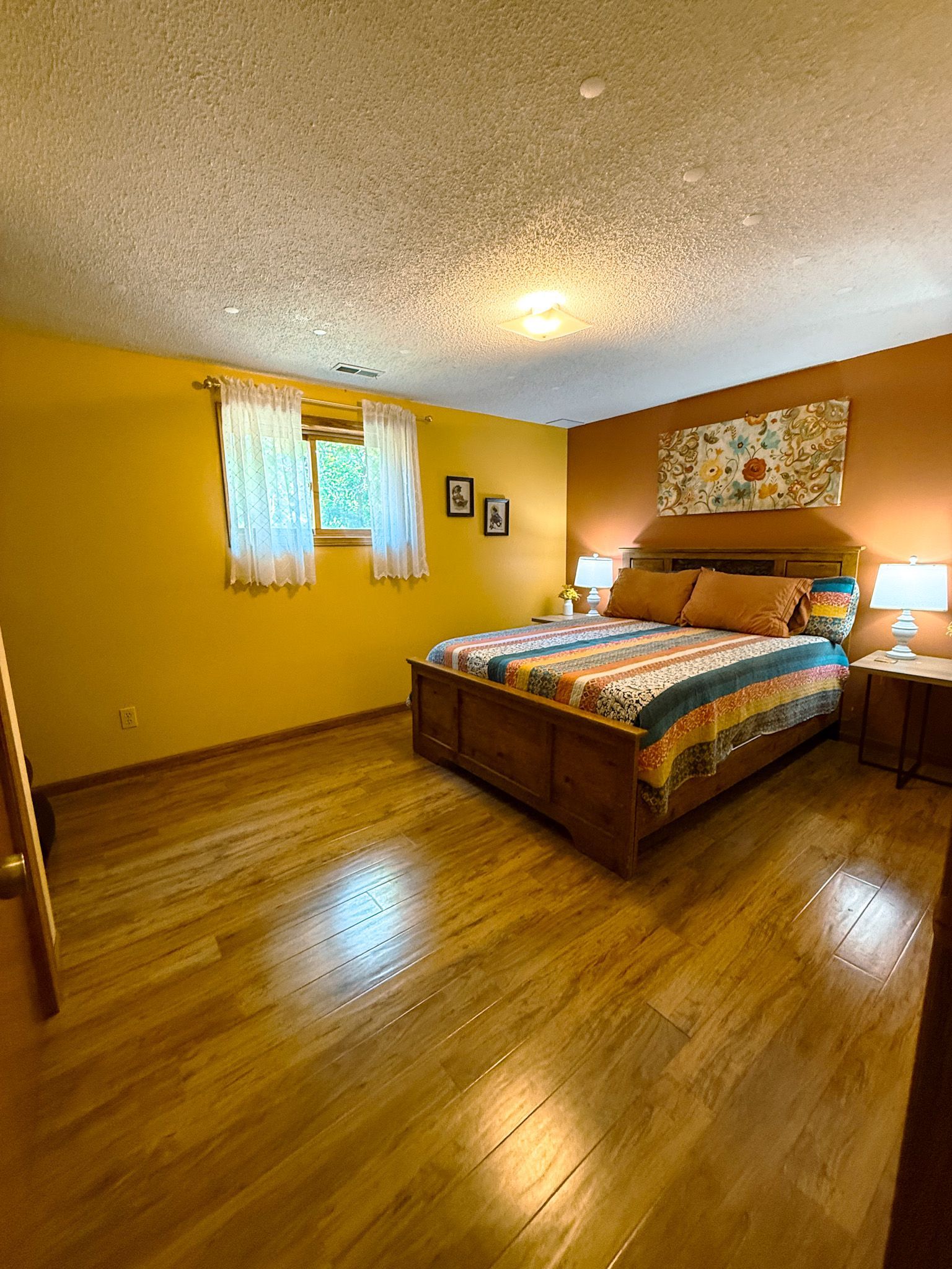 Sunlit bedroom with wood floors, yellow walls, a wooden bed, and a window with sheer curtains