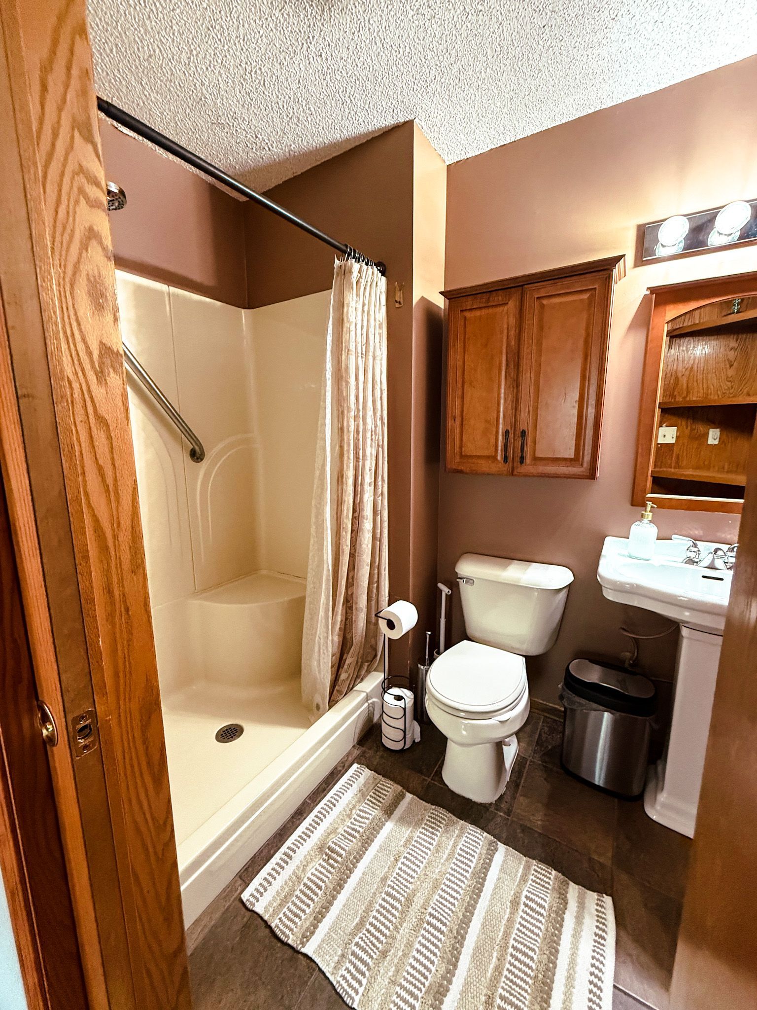 Small bathroom with shower-tub, toilet, vanity, wood cabinets, trash can, and striped rug.