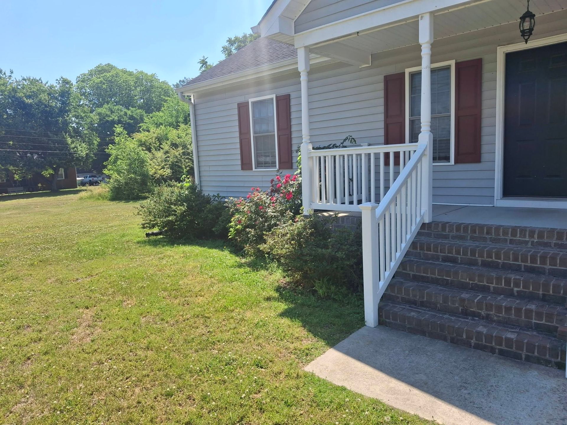 Front porch with white railing, brick steps, and red shutters beside a grassy yard under a sunny sky