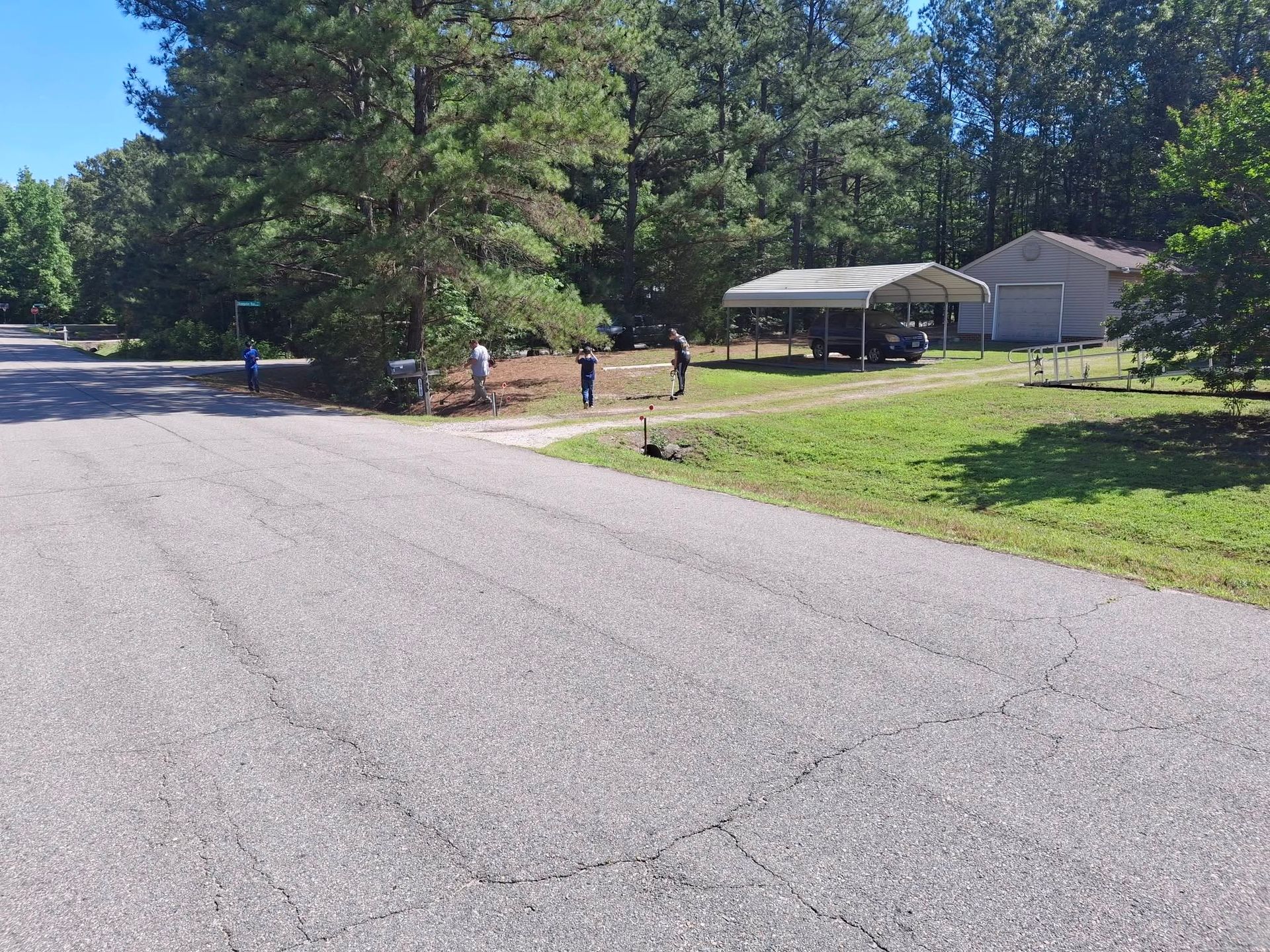 Quiet rural road beside trees, a grassy yard, and a small house with a carport.