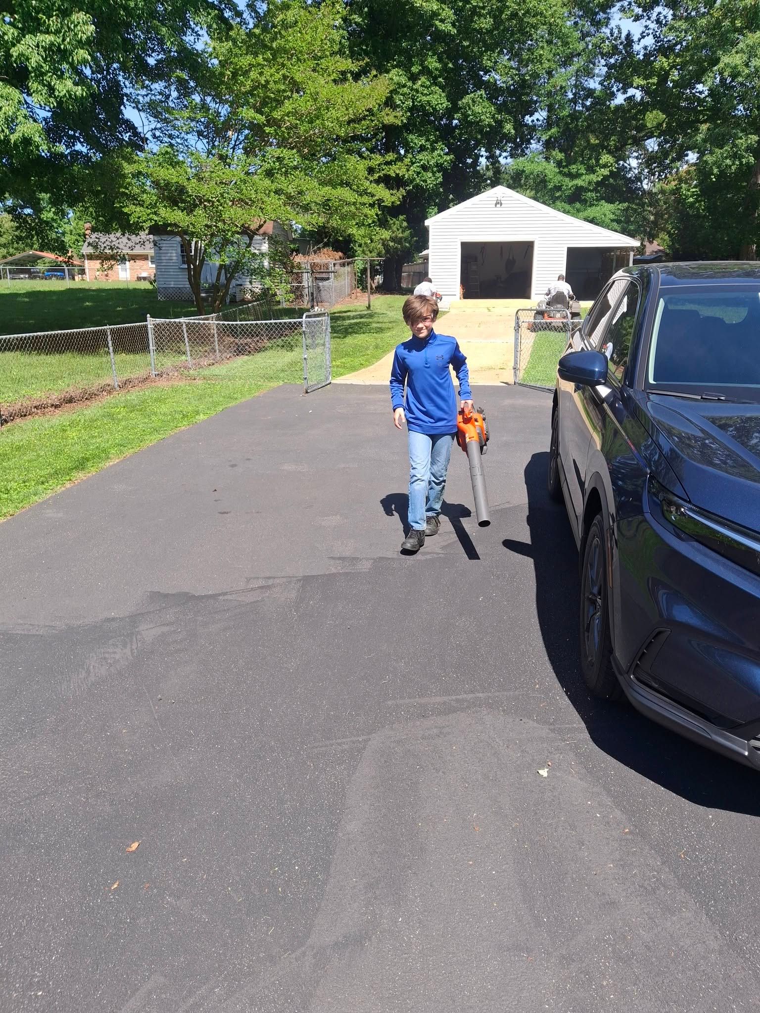 Child in a blue jacket walking a small dog beside a parked black car on a sunny driveway