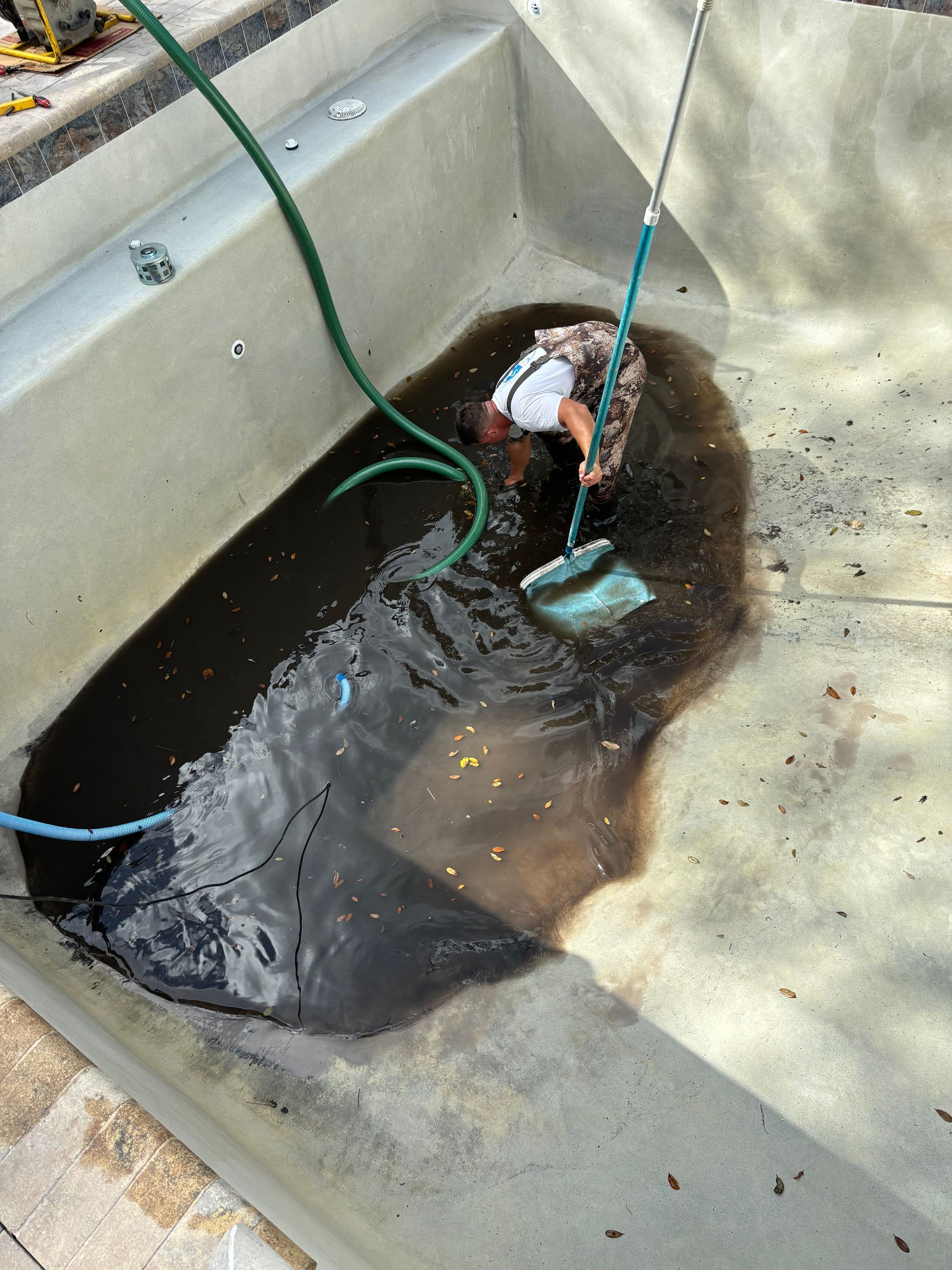 A person standing in the shallow water of an empty pool, using a long-handled net to scoop dark debris from the bottom.