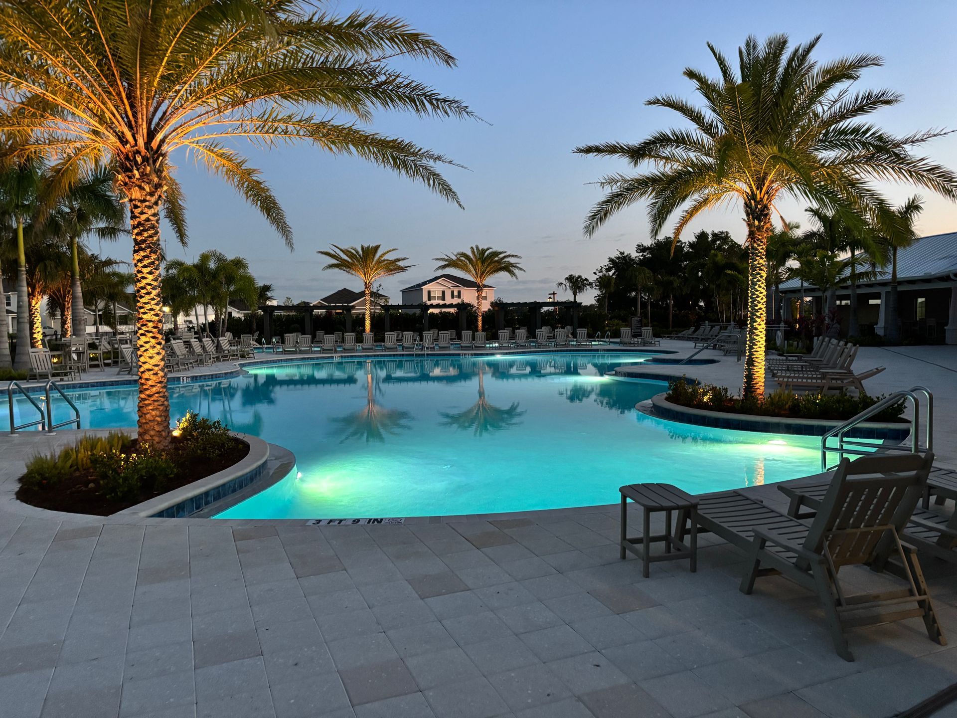 A brightly lit outdoor swimming pool surrounded by palm trees and patio chairs during twilight.