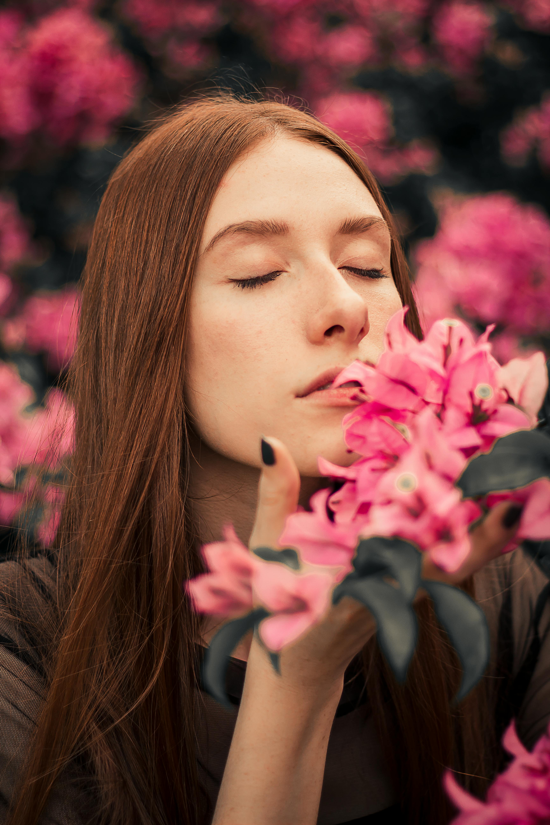 Woman with red hair smelling pink flowers, eyes closed, in a garden.
