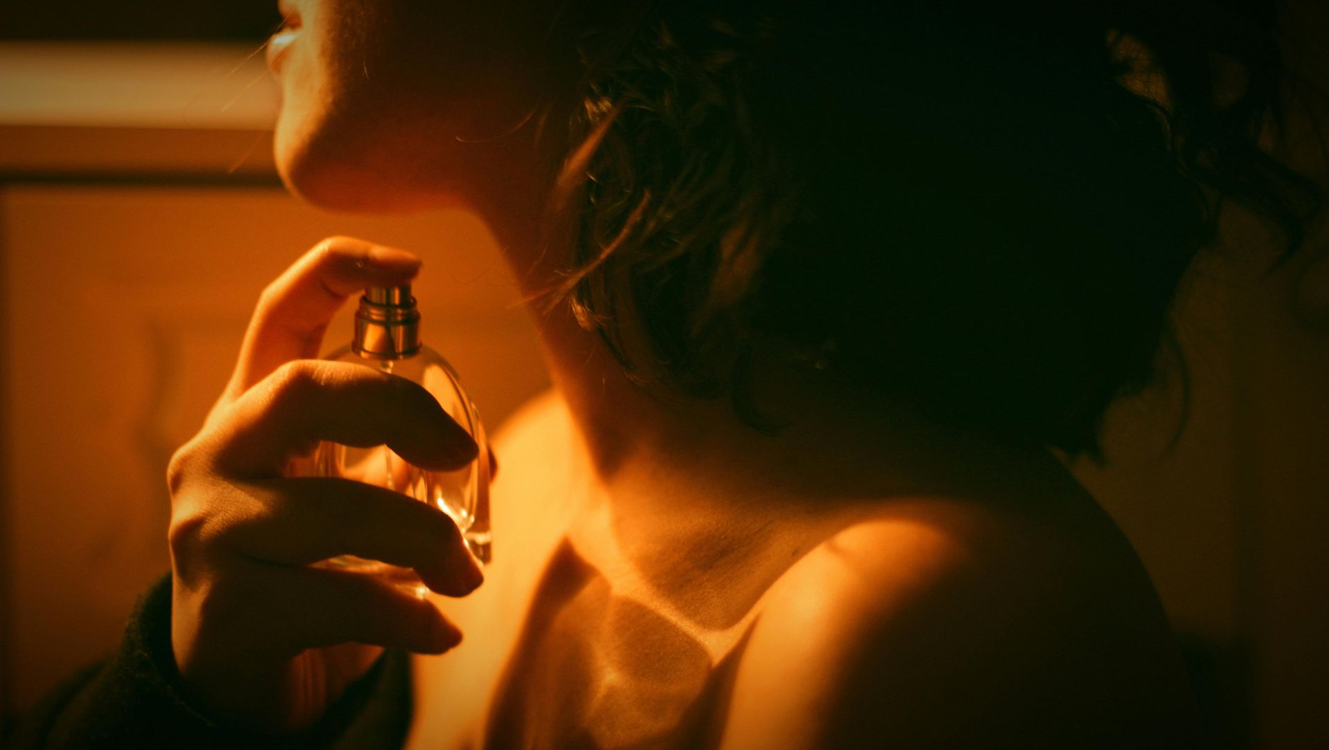 Vrouw spuit parfum op haar hals. Warm licht, focus op het flesje en de hand.