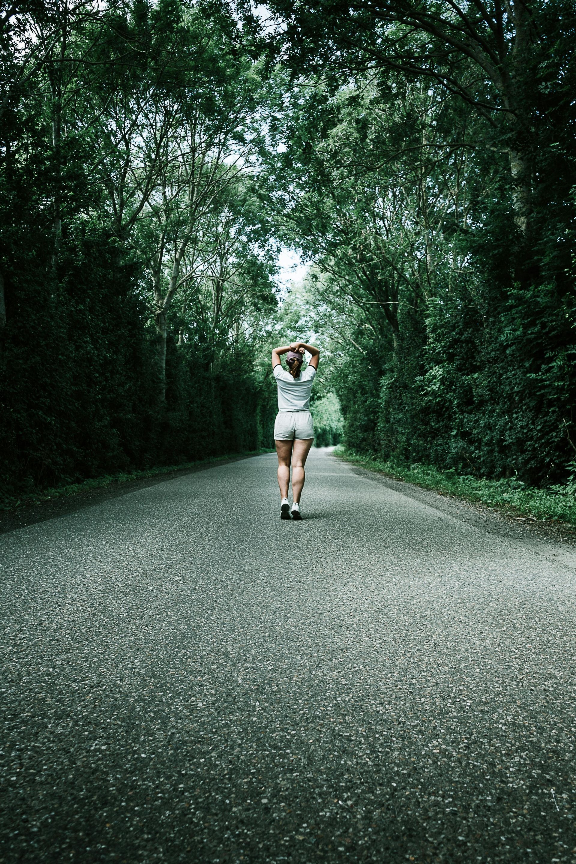 Woman walking down a paved path lined by trees, arms raised.