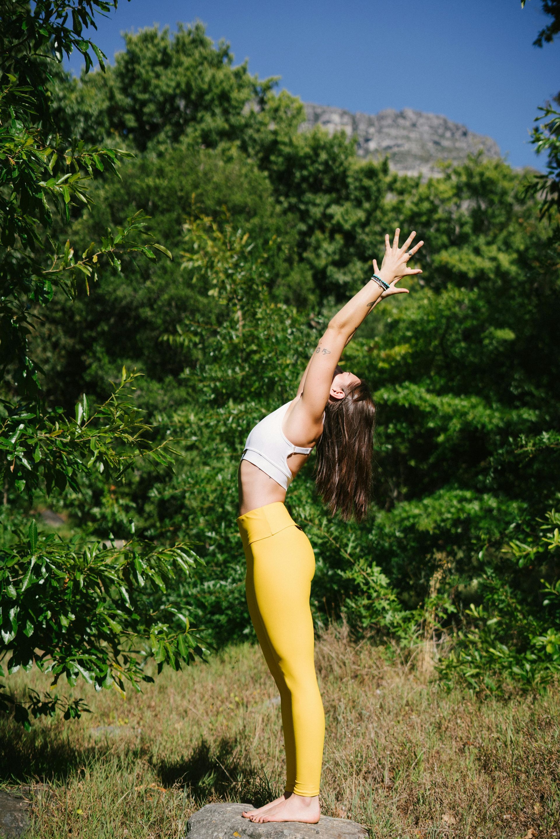 Woman in a white top and yellow leggings, arching back in yoga pose outdoors, arms raised.
