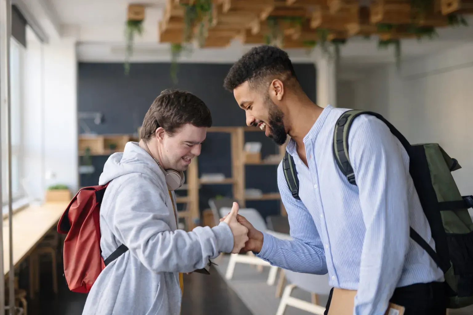 Support worker and NDIS participant with Down syndrome shake hands in a bright indoor space, one with a red backpack, both smiling.